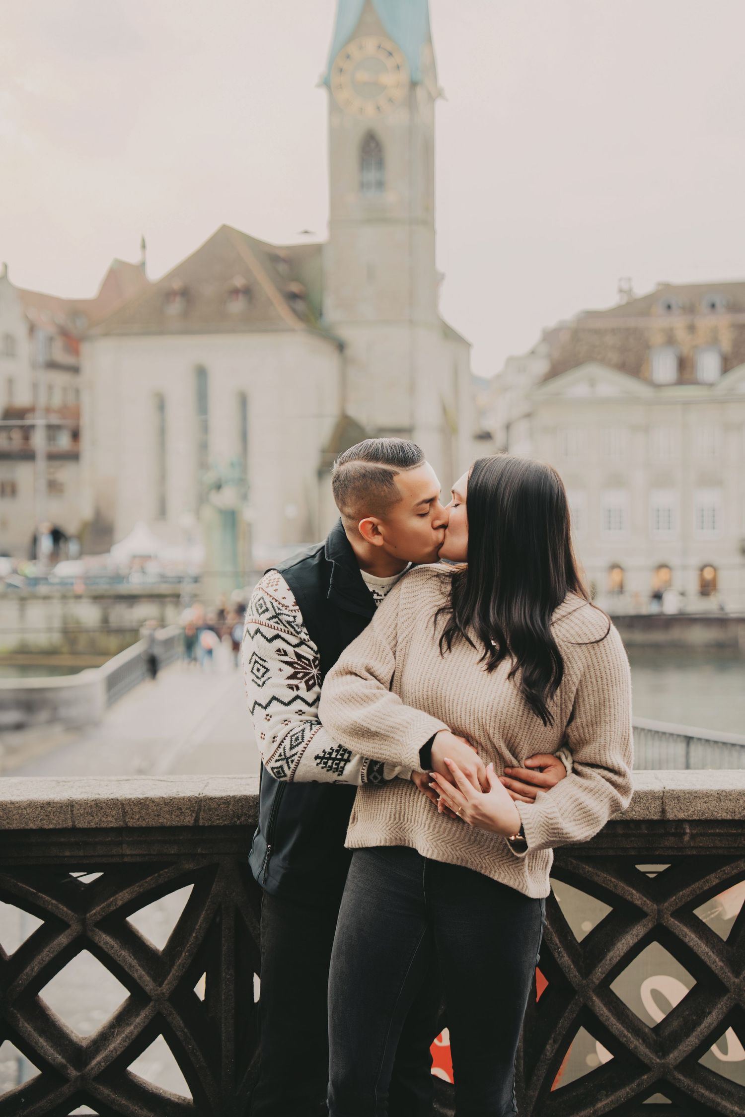 Couple sharing a kiss on a bridge with Fraumunster Church and Zurich cityscape in background.