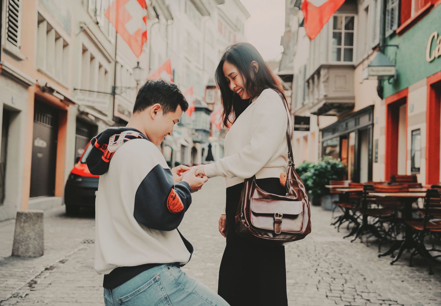 Man kneels on cobblestone street proposing to smiling woman in European old town setting.