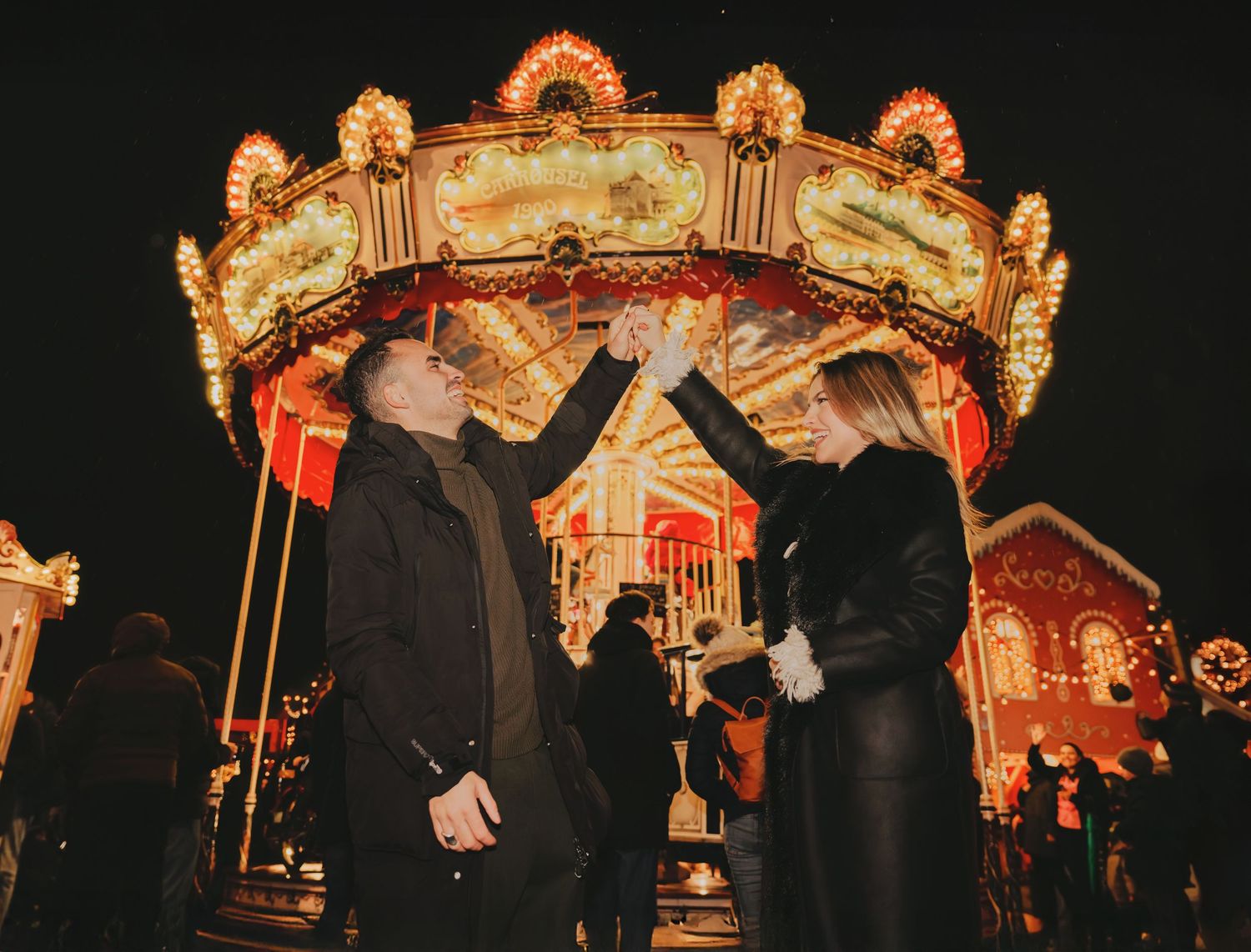 Couple toasting drinks in front of a beautifully lit carousel at a festive Christmas market at night.