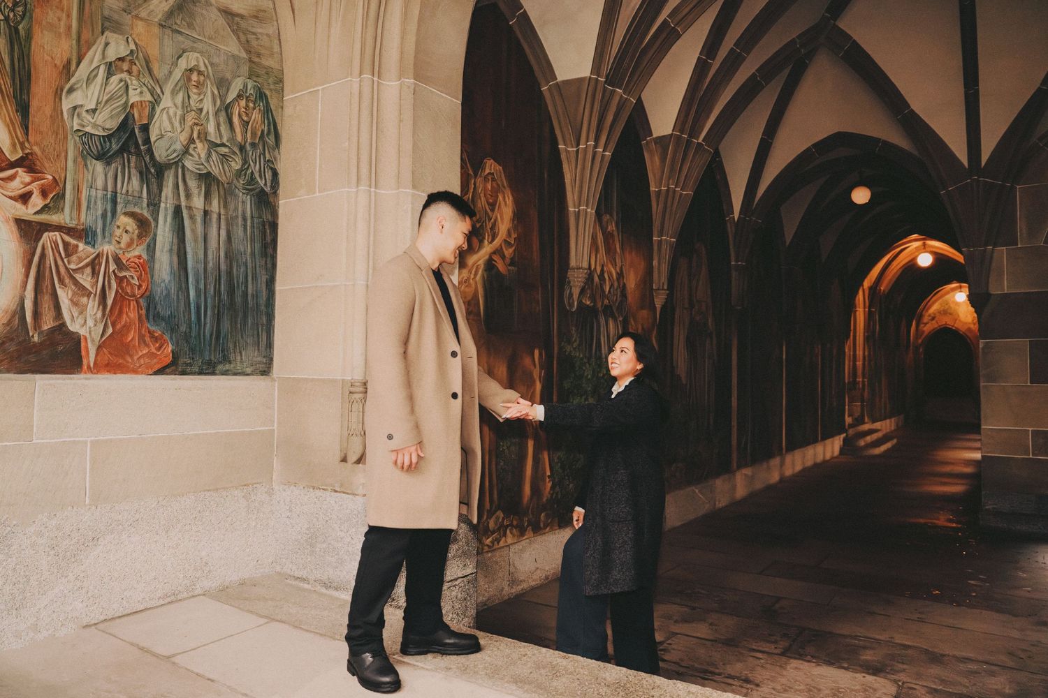 Couple holding hands in a Gothic stone corridor with arched ceilings and painted murals.