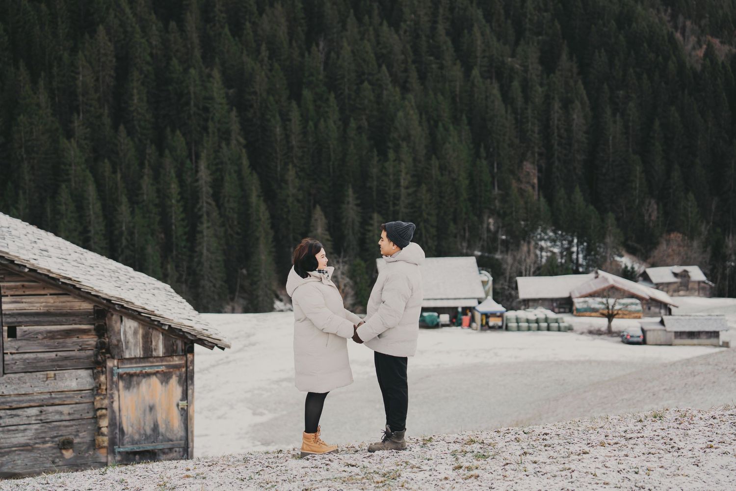 Couple holding hands in snowy alpine village with rustic wooden cabin and dense forest backdrop.
