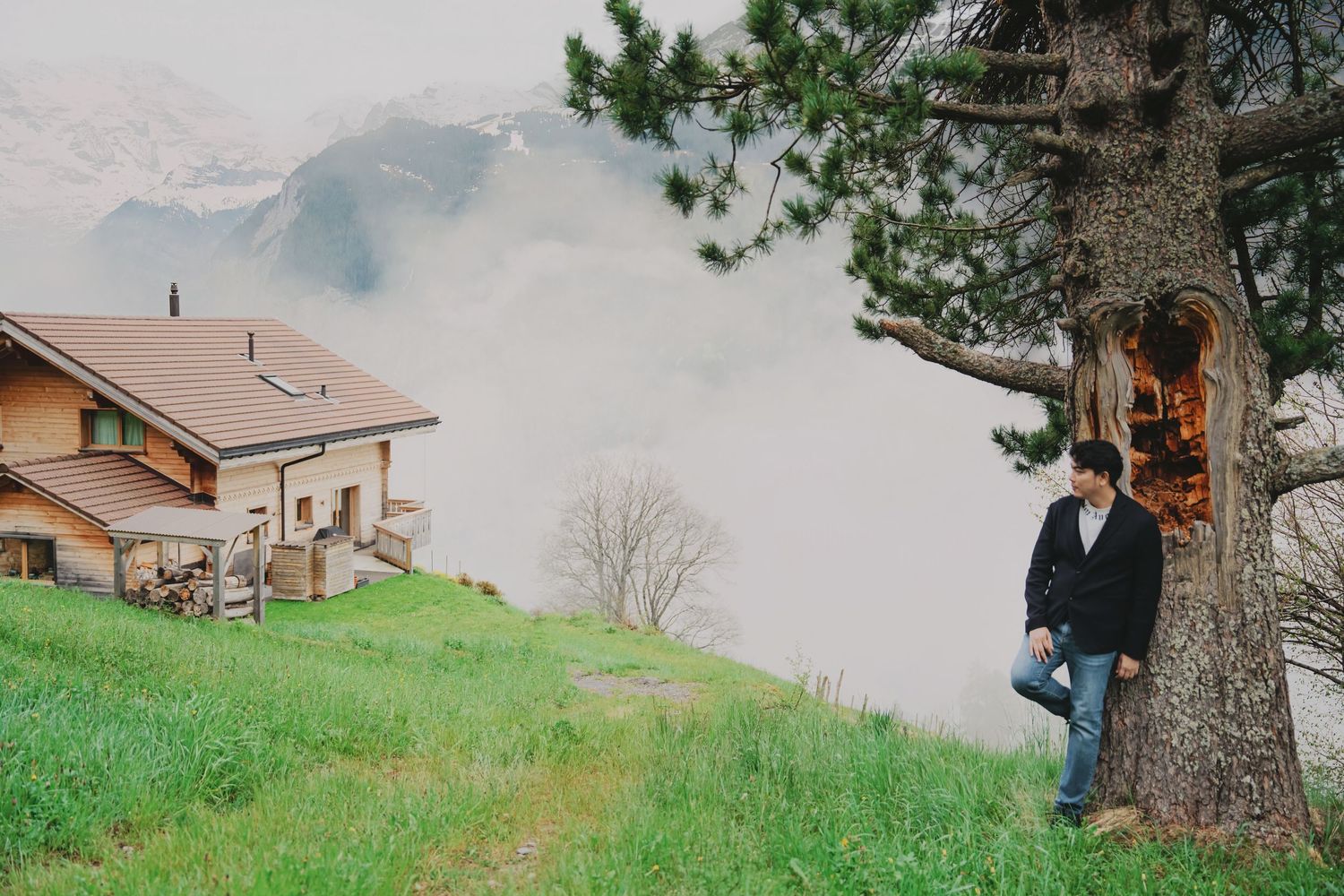 Man leaning against a large pine tree on a misty green hillside beside a Swiss alpine chalet.