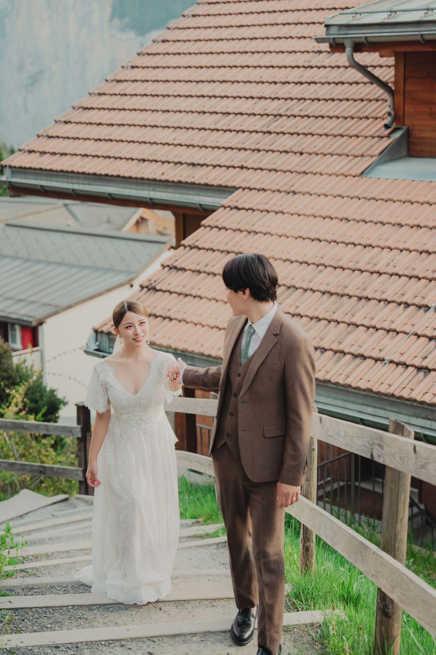 Smiling bride and groom in brown suit walking down wooden steps with red-roofed alpine buildings.