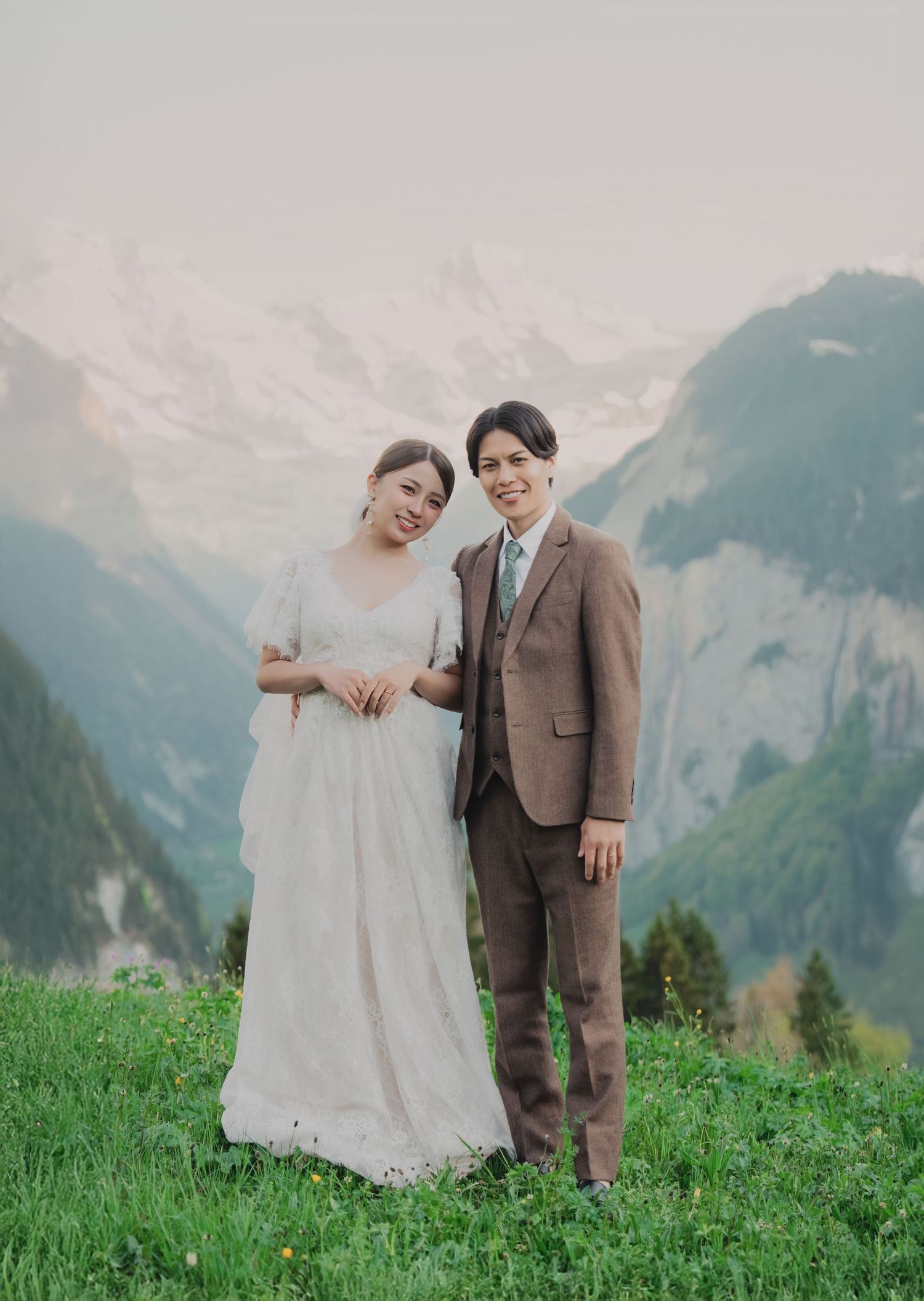 Smiling Asian couple in wedding attire posing together on green alpine meadow with misty Swiss mountains behind.