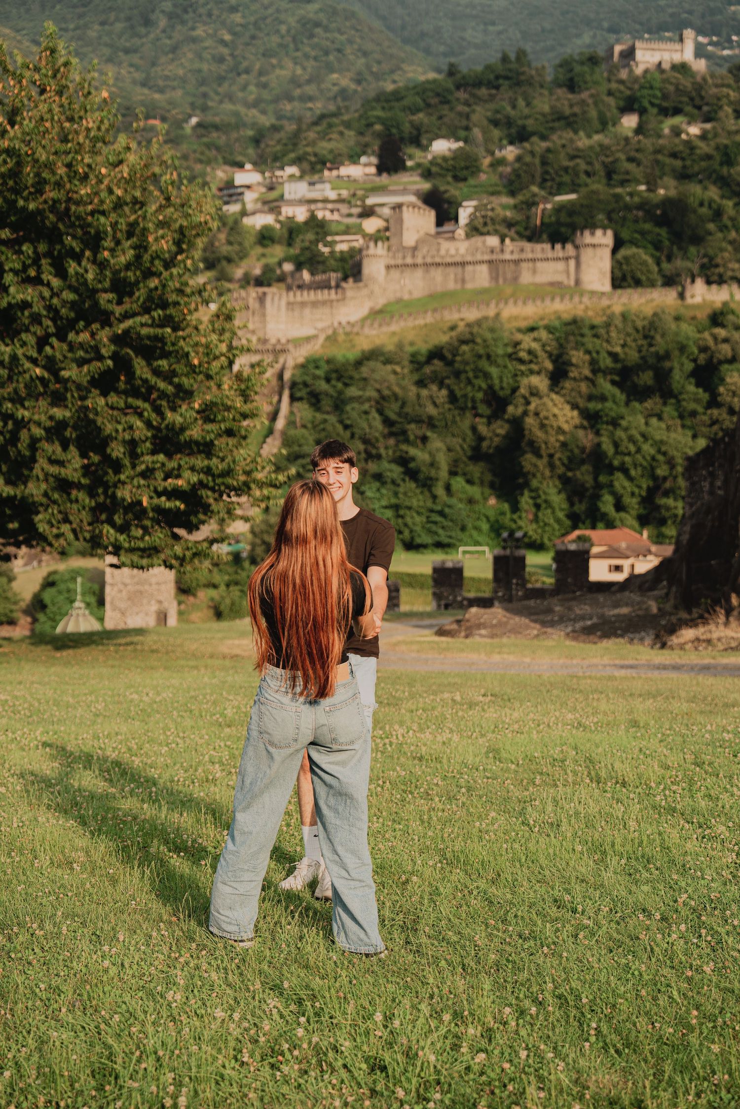 Romantic couple sharing a kiss on a grassy lawn with a medieval castle wall backdrop.