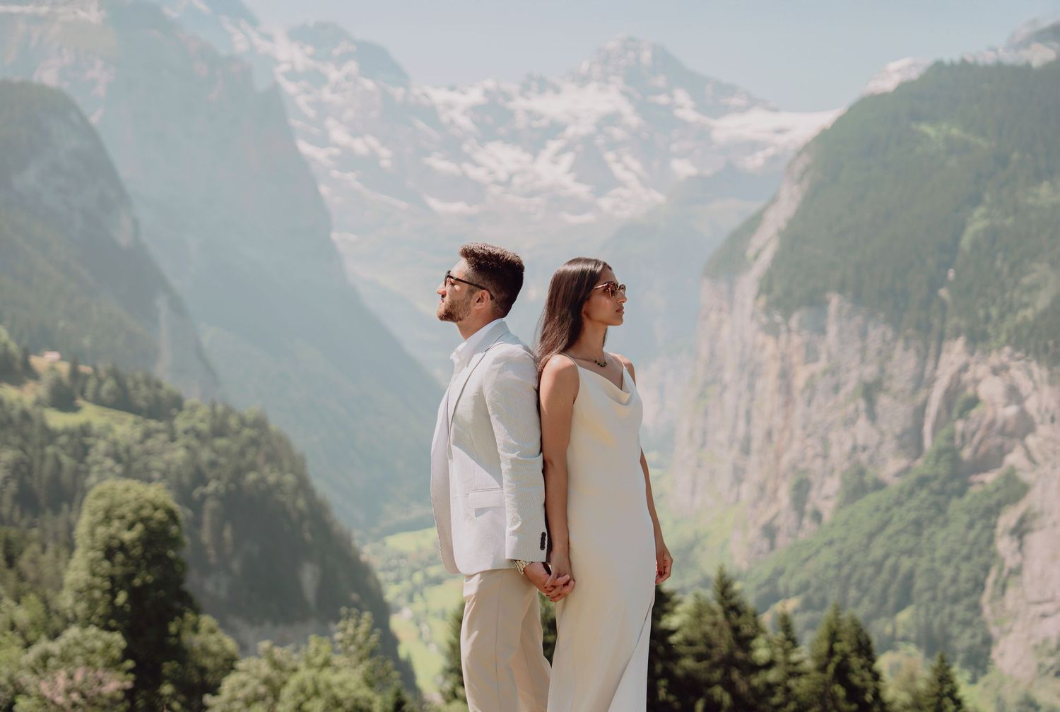 Stylish couple standing back-to-back on a scenic alpine overlook with snow-capped Swiss mountain peaks.