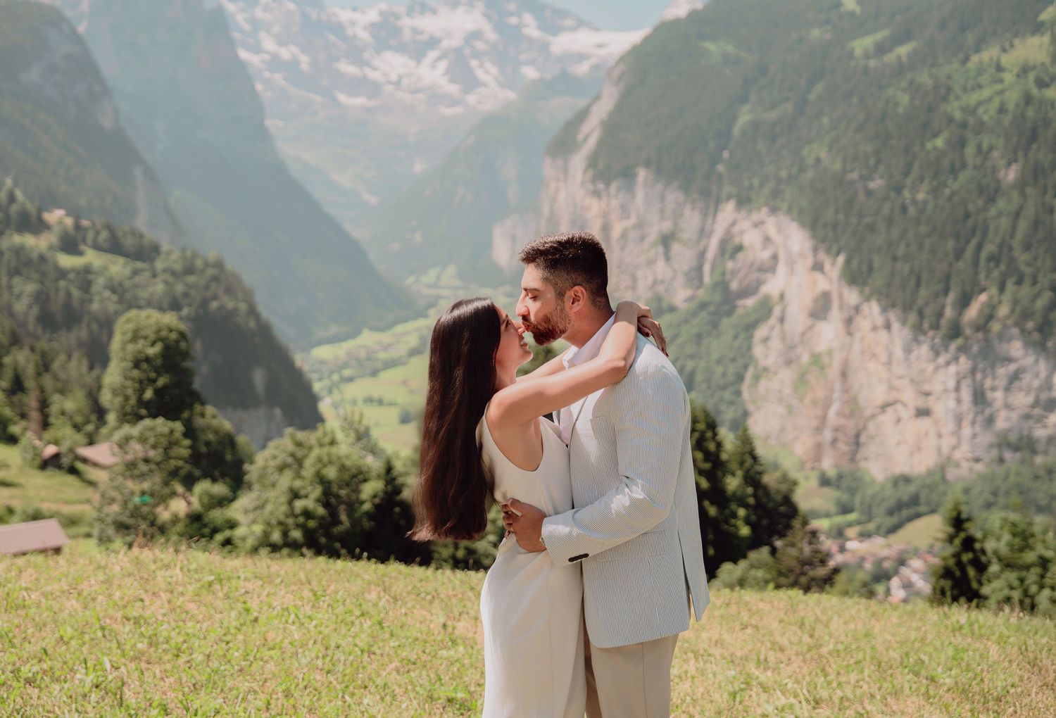 Couple shares a tender kiss in a sunlit Alpine meadow with dramatic Swiss mountain valley views behind them.