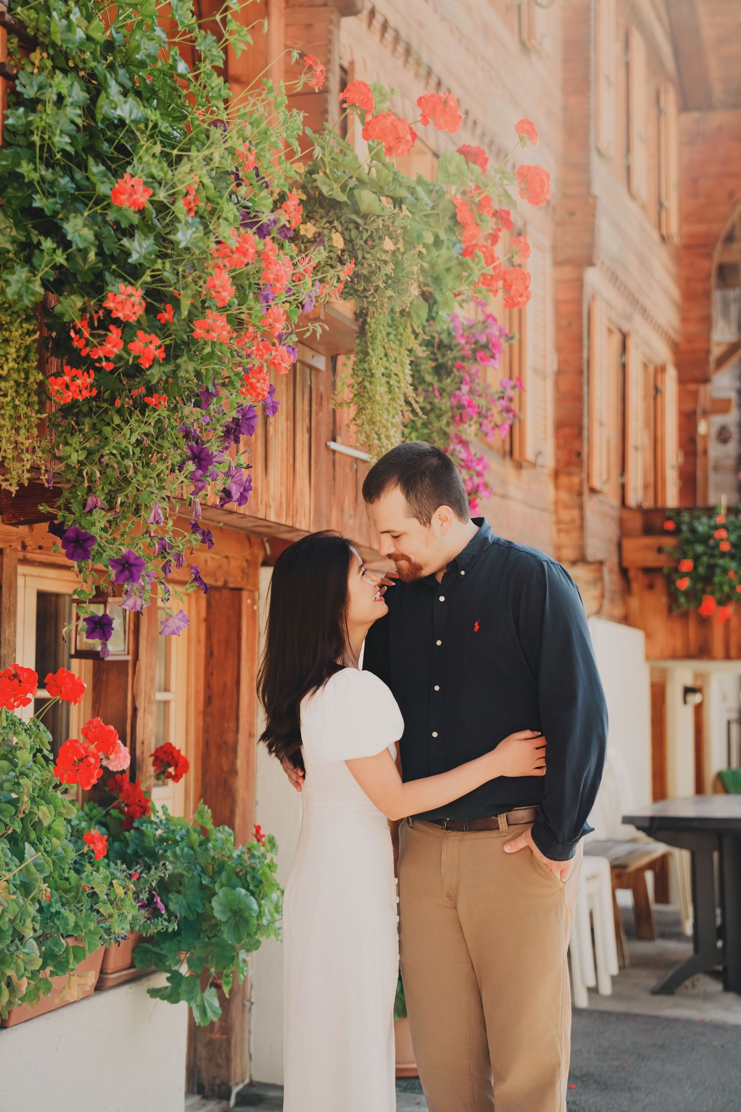 Romantic couple sharing a tender moment surrounded by vibrant red and purple hanging flowers on a rustic wooden building.