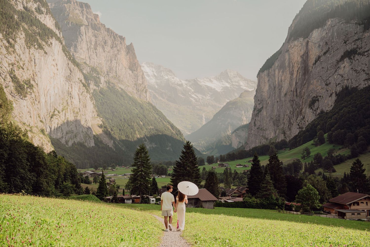 Couple walking hand-in-hand with white parasol through green Swiss Alps valley meadow.