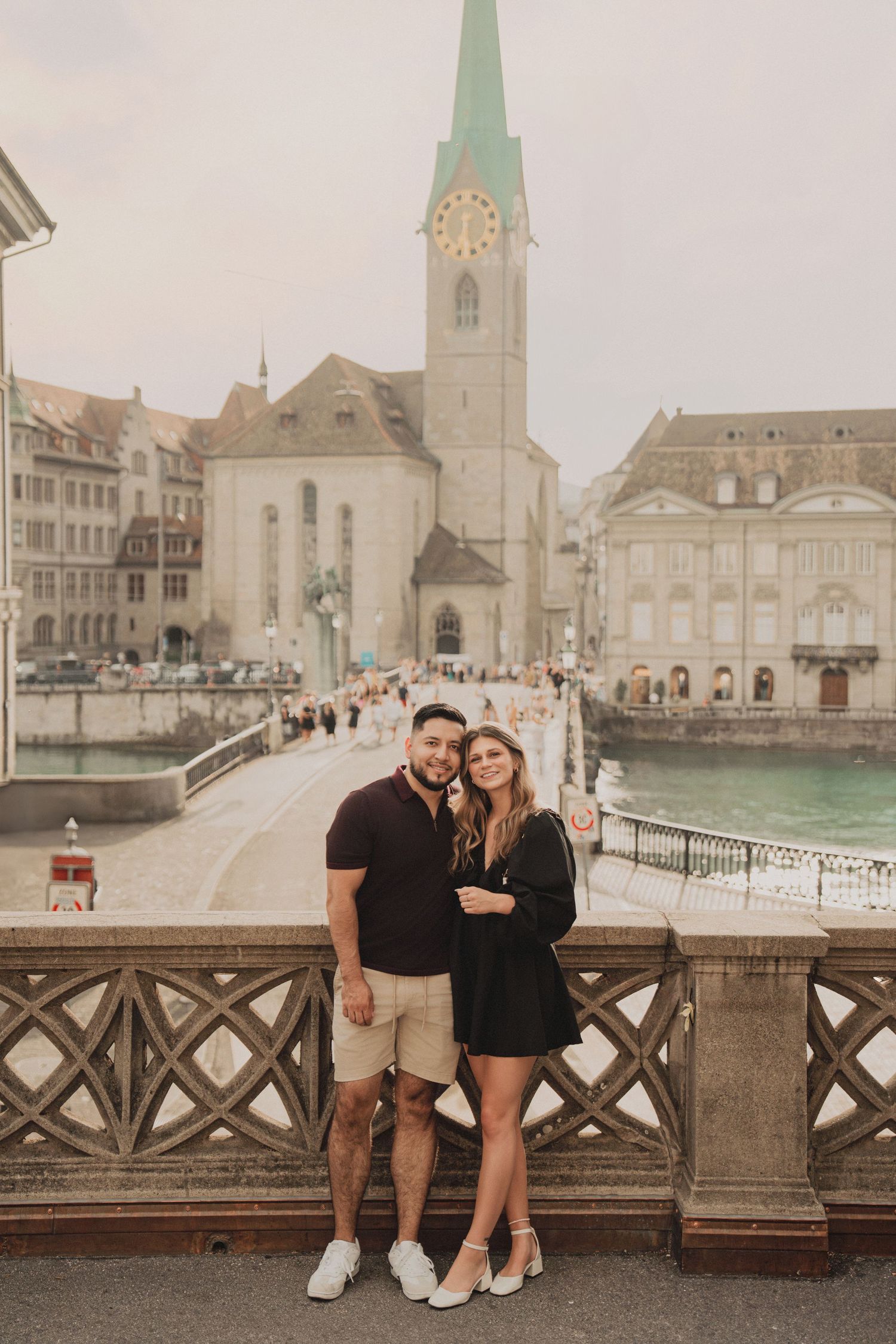 Couple posing on Zurich bridge with Fraumunster church clock tower and river in background.