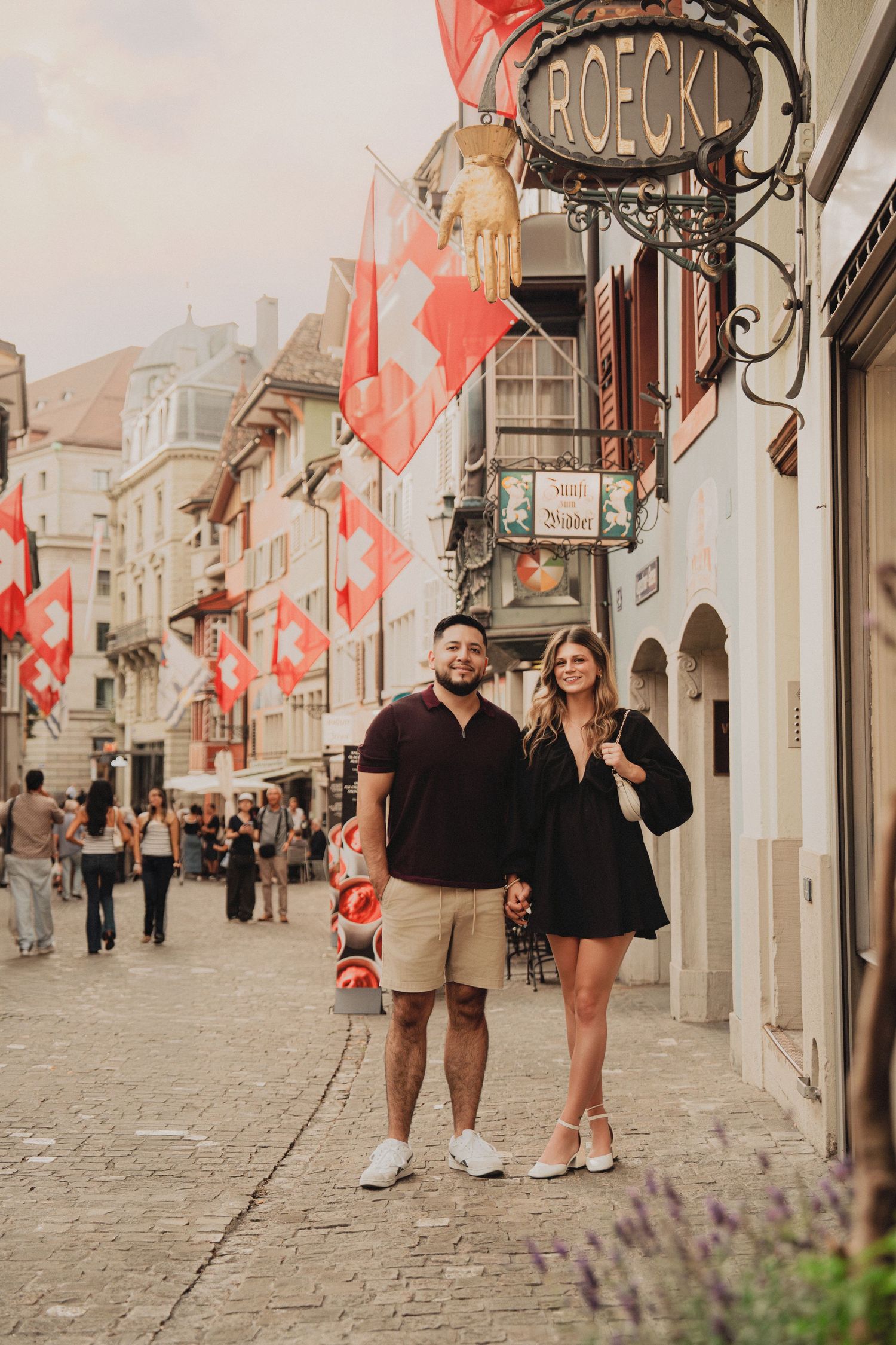 Couple holding hands on a cobblestone street in Zurich, Switzerland, with Swiss flags and the Roeckl shop sign behind them.