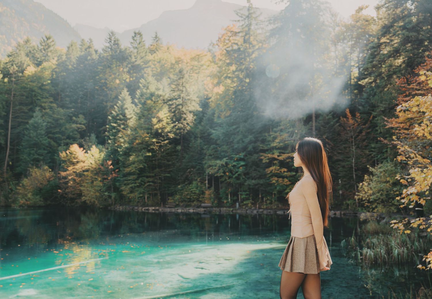 Young woman in beige outfit stands by a misty turquoise alpine lake surrounded by autumn forest.