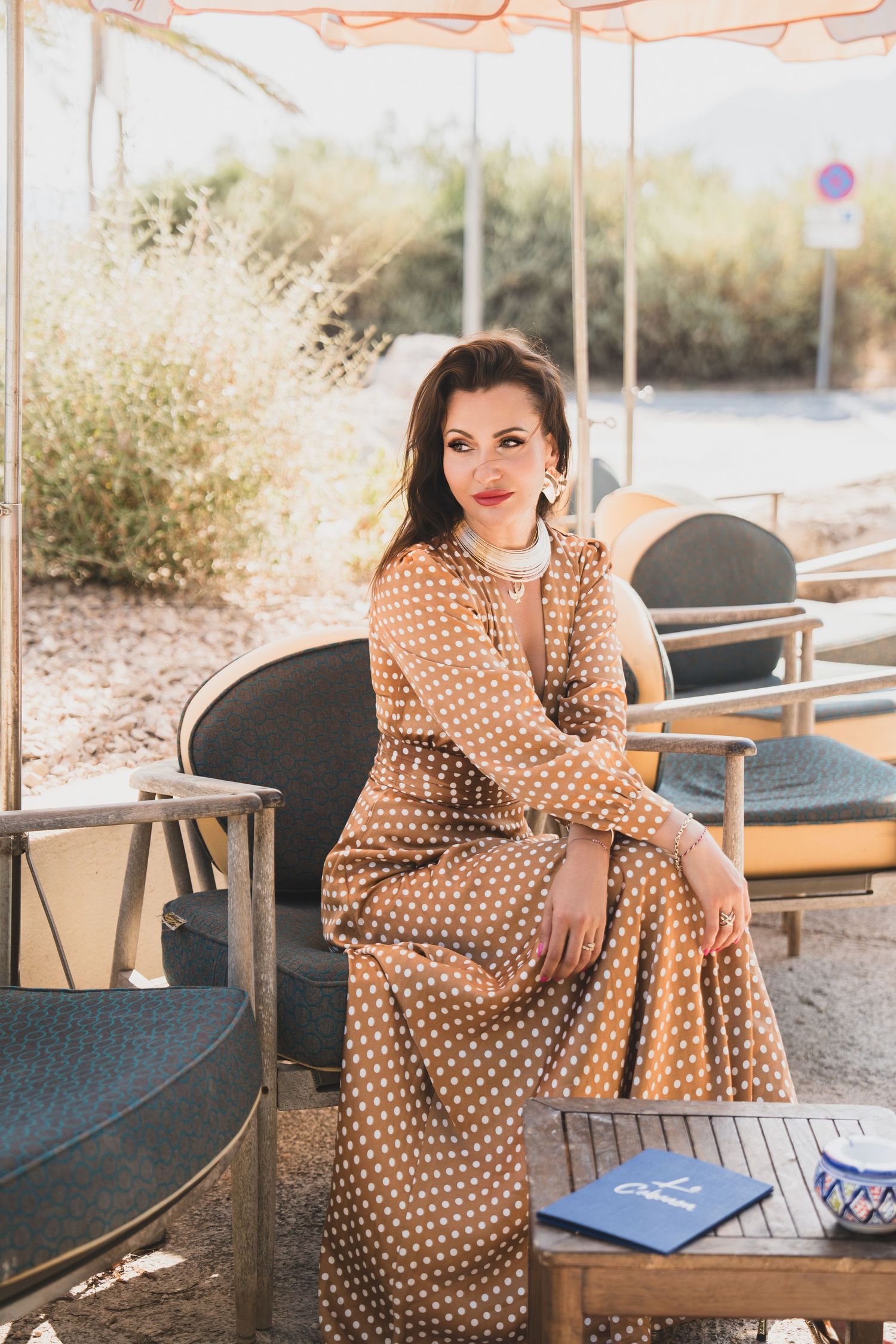 Woman in brown polka dot maxi dress sitting at outdoor cafe table in sunny Mediterranean setting.