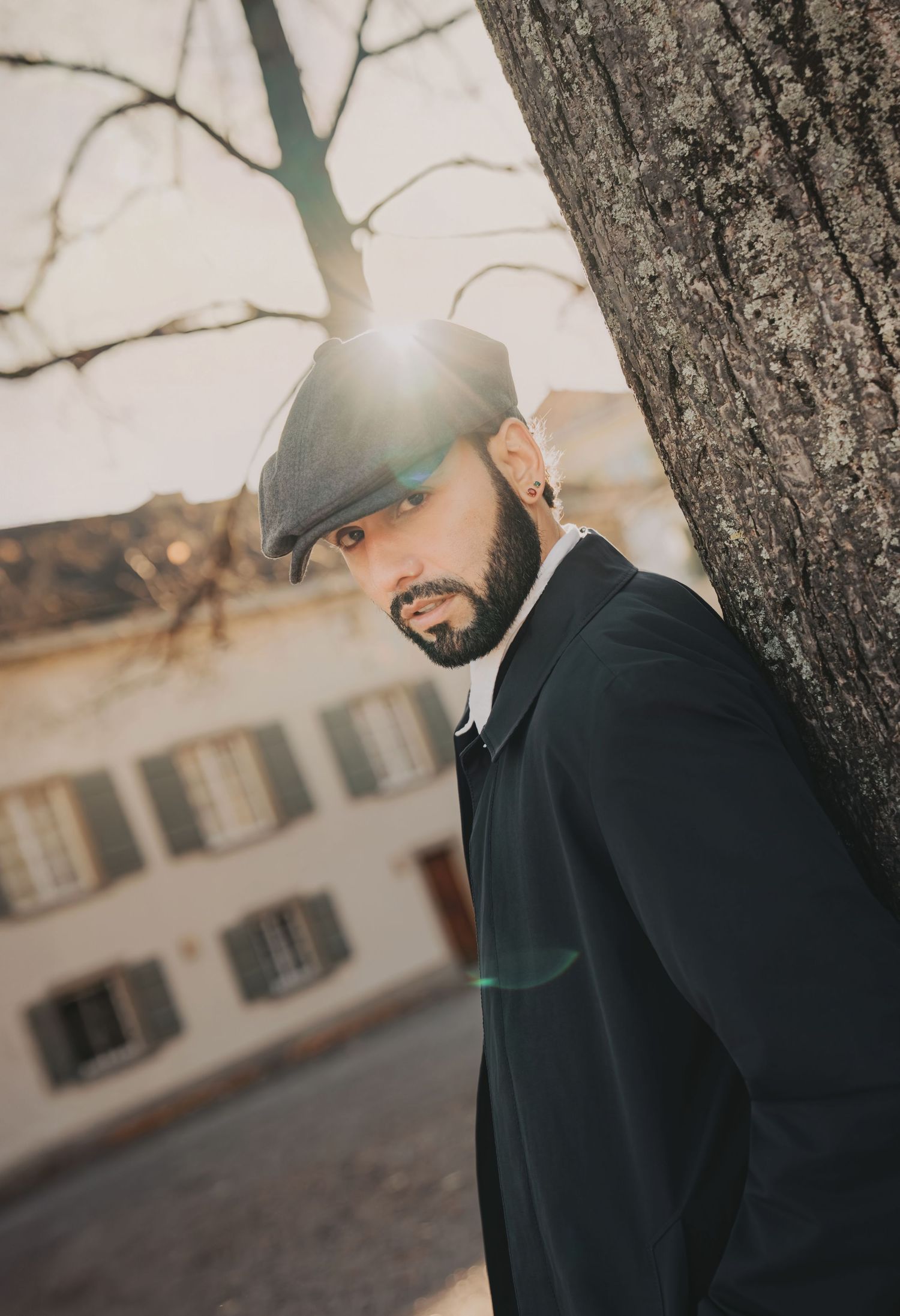 Bearded man in black coat and flat cap leaning against a tree with sunlight flaring behind him.