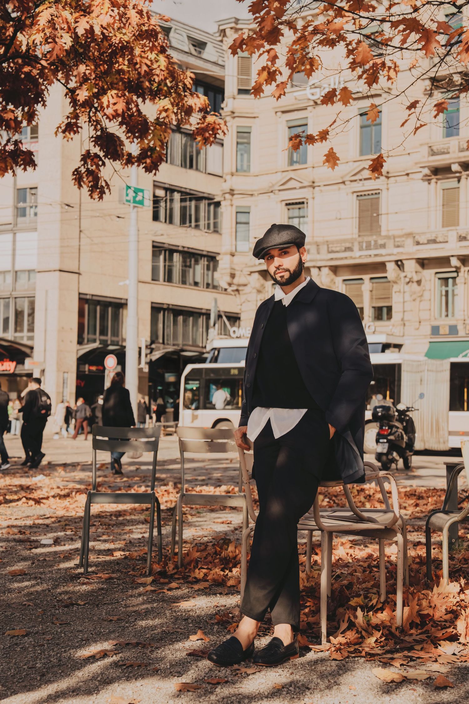 Fashionable man in black coat and flat cap standing among autumn leaves in urban plaza.