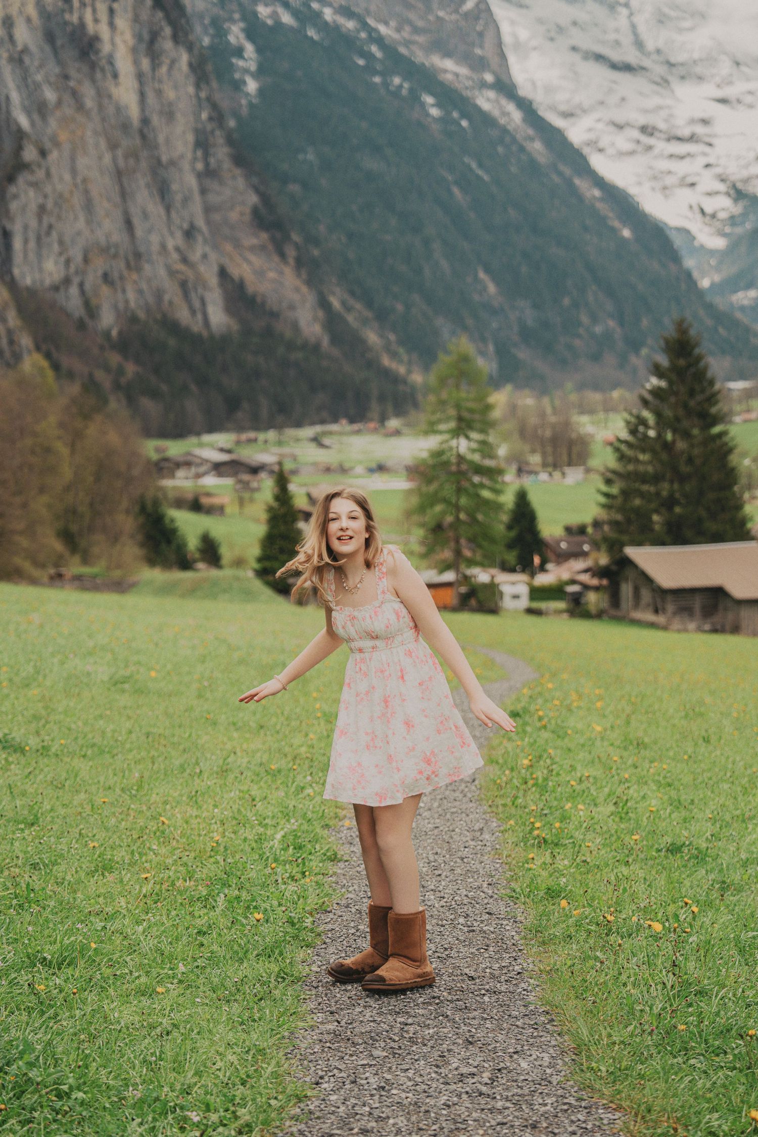 Joyful young woman in floral dress and brown boots twirling on a path in a stunning Swiss Alpine valley.