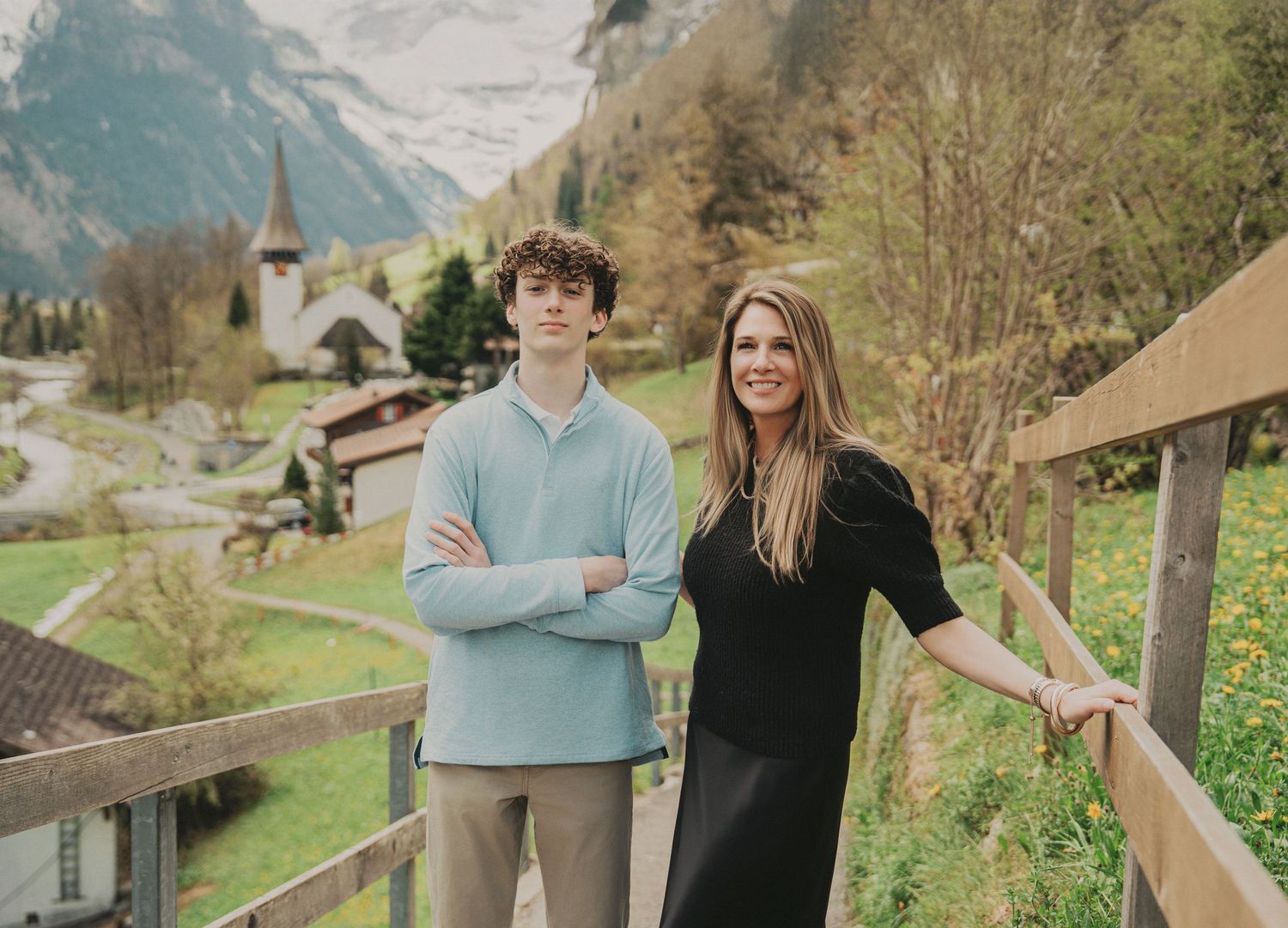 A young man and woman pose on a wooden balcony overlooking a scenic Swiss alpine village with mountains.