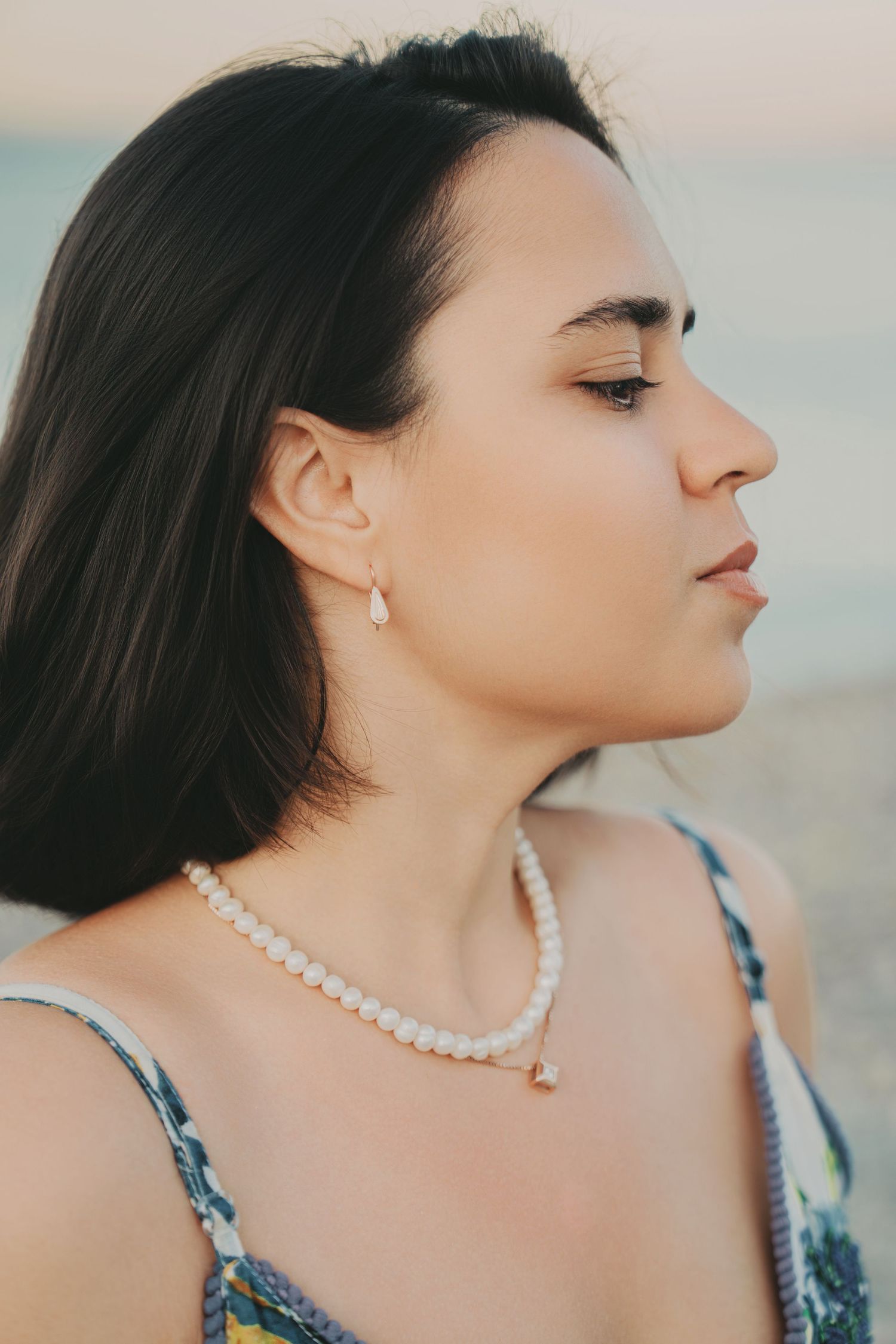 Close-up profile portrait of a young brunette woman wearing pearl necklace and drop earrings at beach.