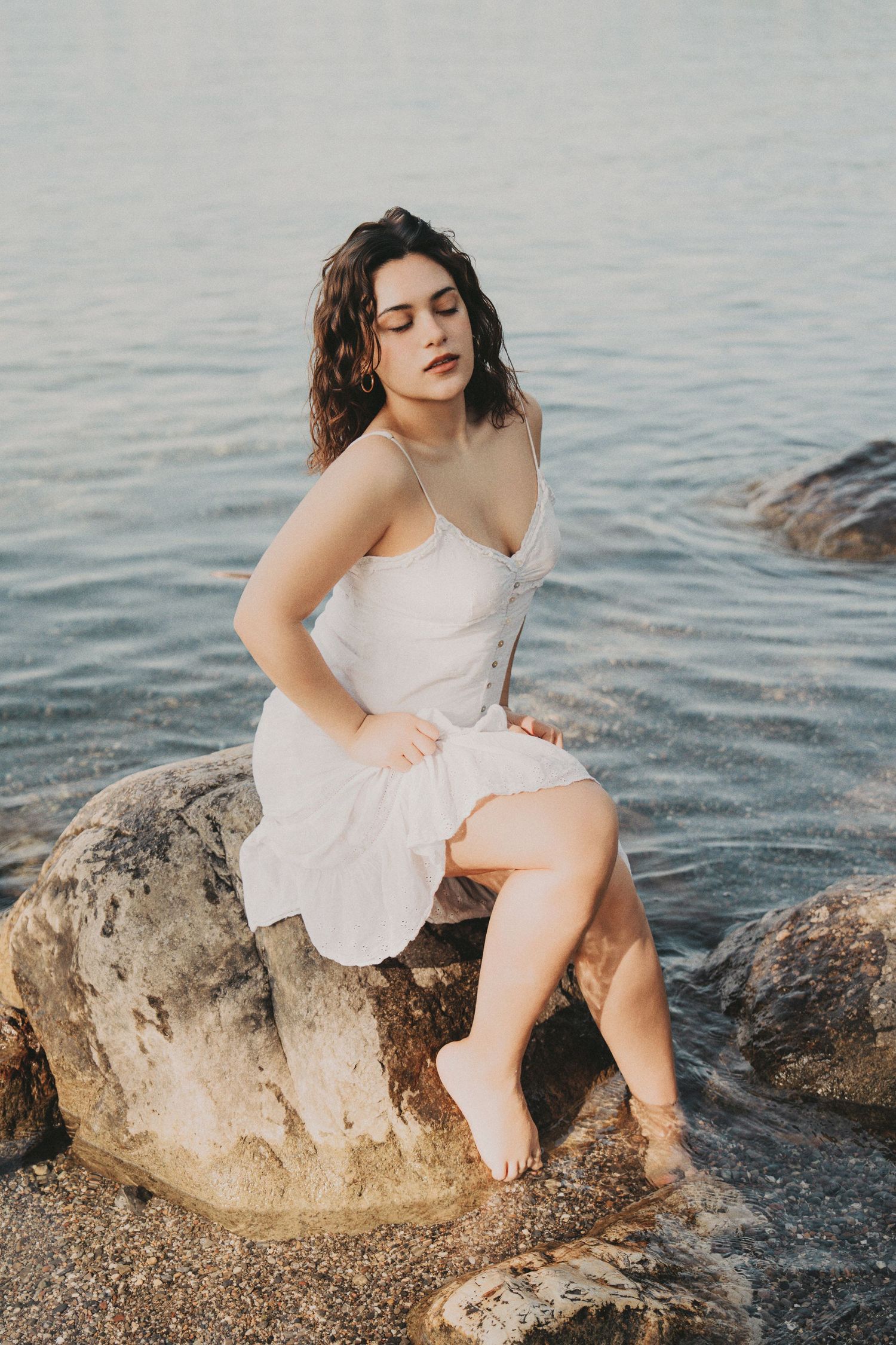 Woman in white dress sitting barefoot on rocks with feet in shallow water.