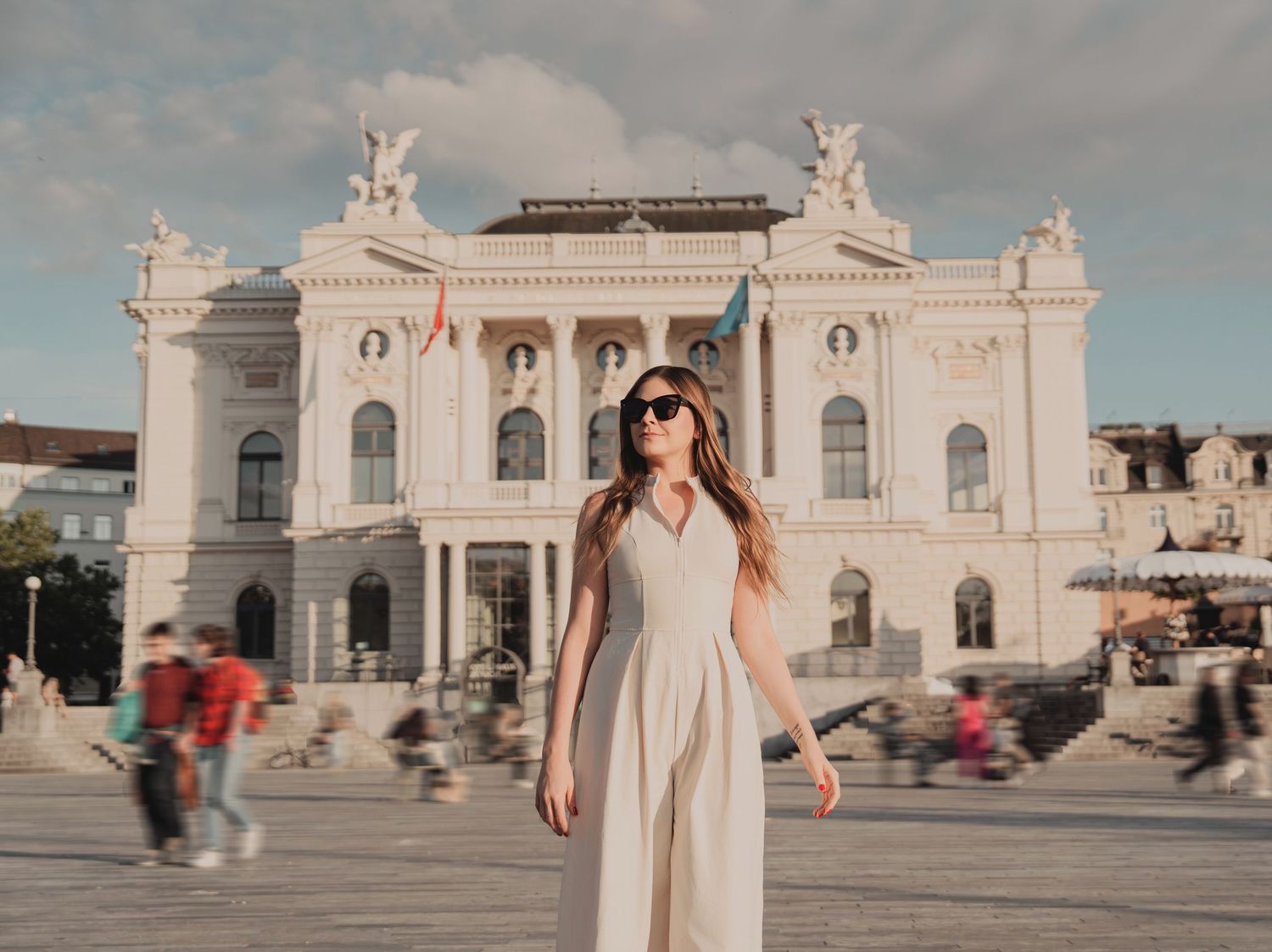 Woman in white dress standing before ornate white neoclassical opera house in city square.