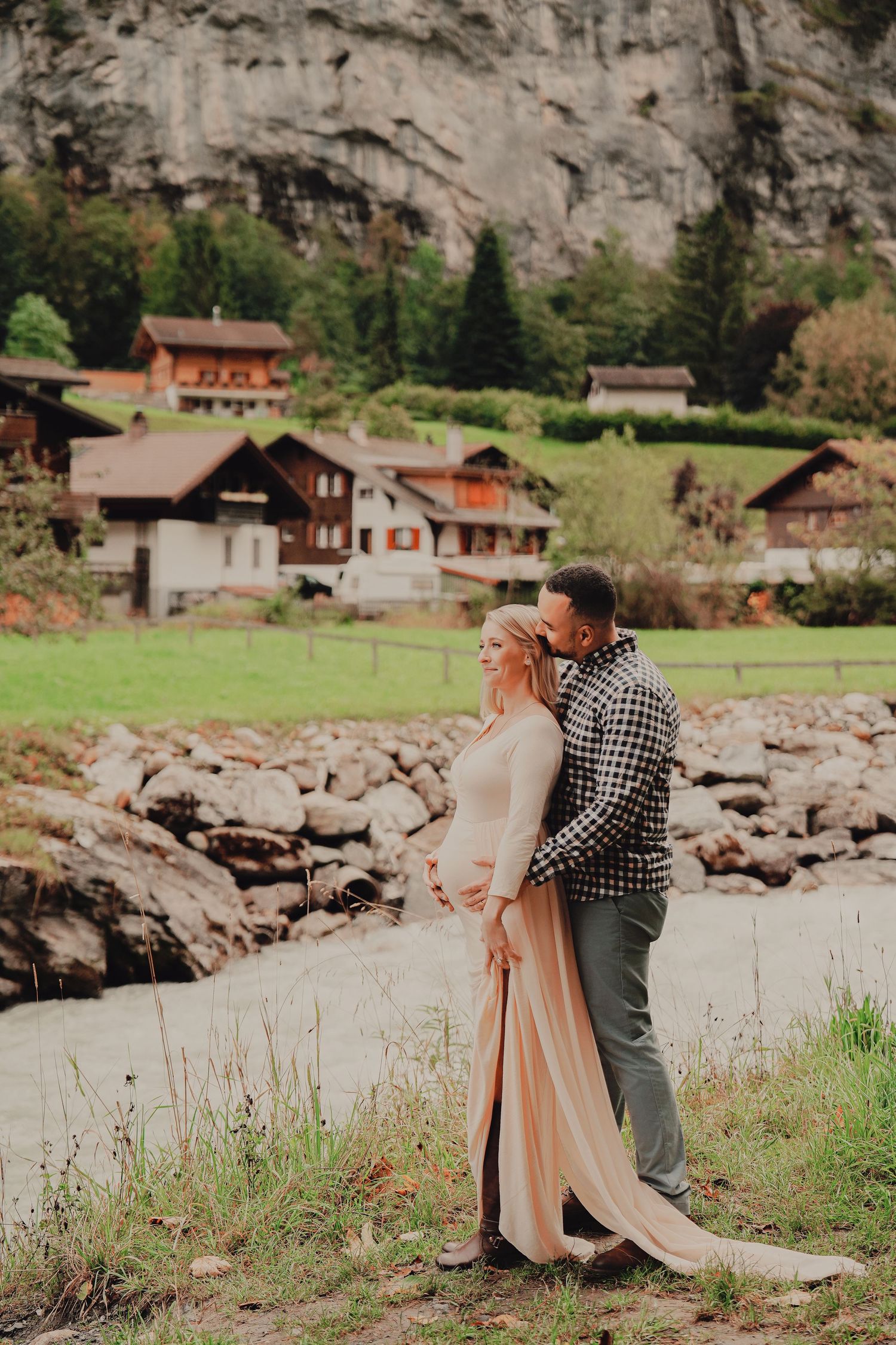 Couple embracing near rocky riverbank with traditional Swiss chalets and green mountains behind.