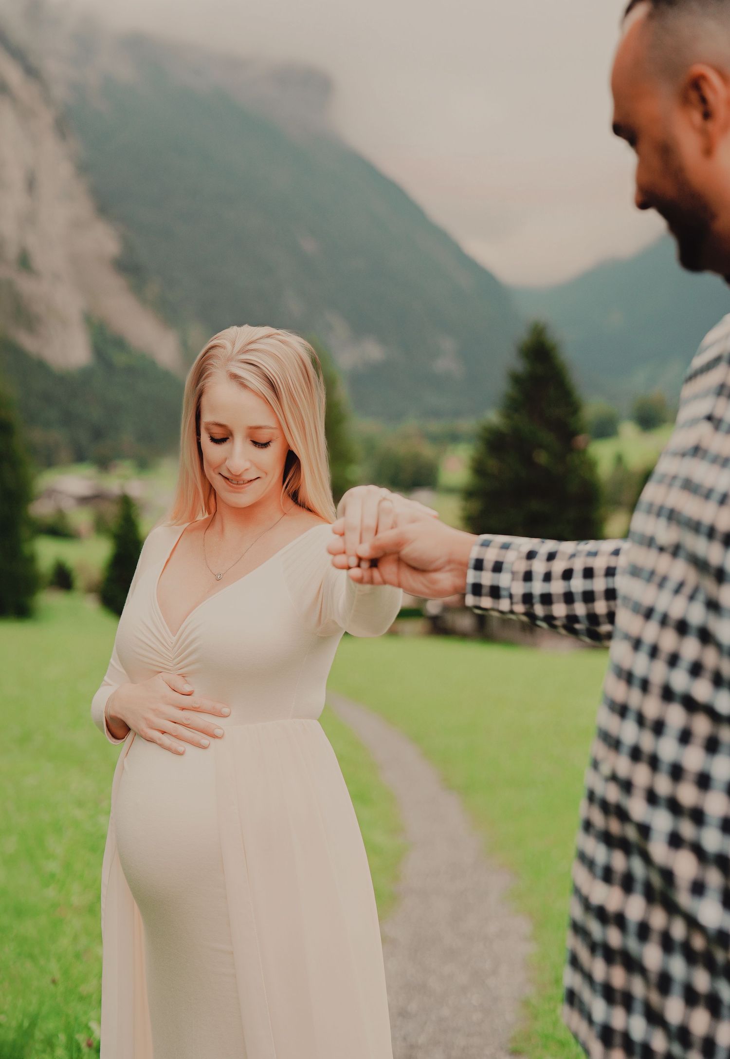 Expectant mother in cream dress tenderly holds belly while partner holds her hand in Alpine meadow.