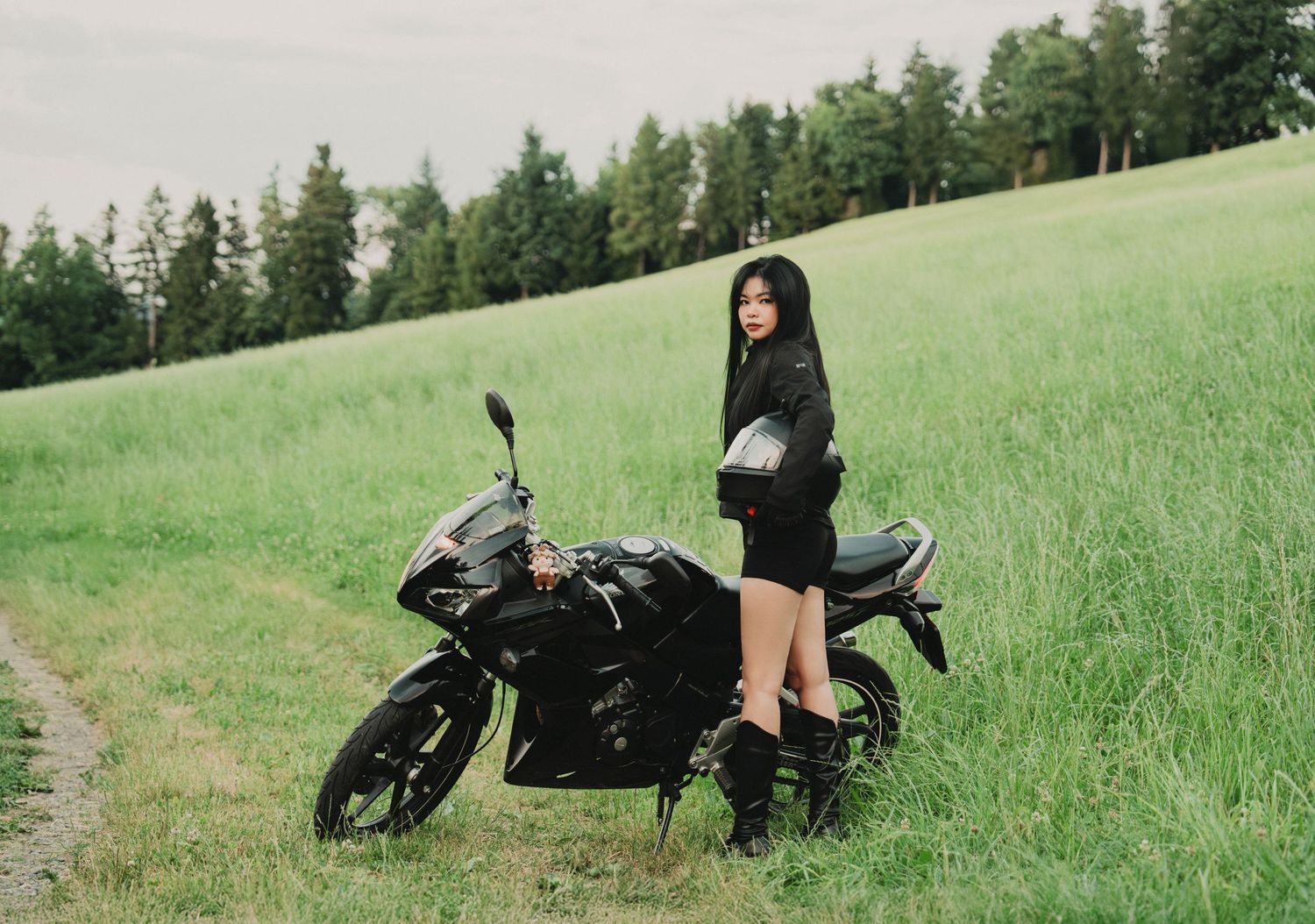Young Asian woman in black outfit standing beside black motorcycle on rural grassy hillside.