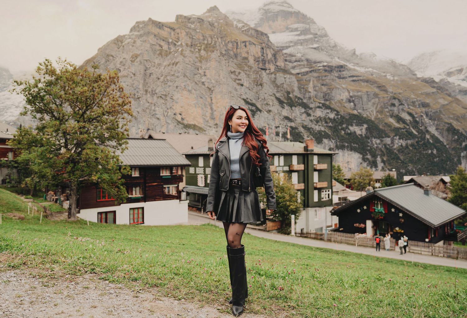 Stylish woman in black leather outfit posing with dramatic mountain peaks and Swiss village behind her.