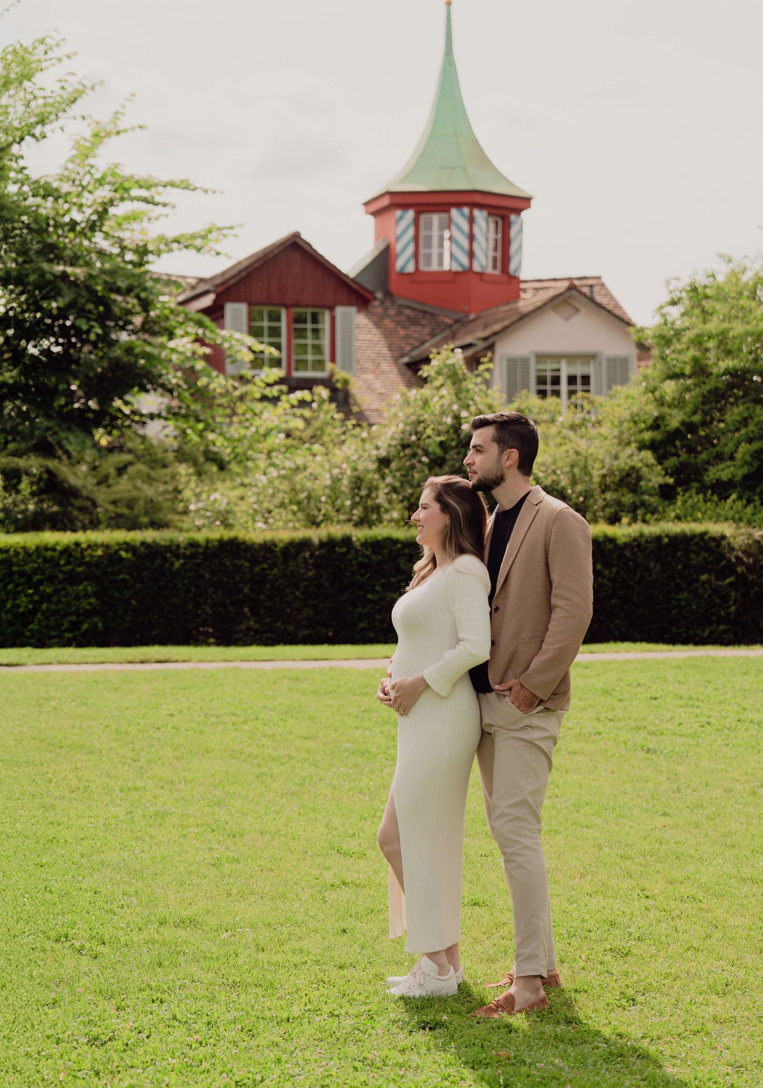 Pregnant couple standing together on green lawn with a picturesque red-spired building behind them.
