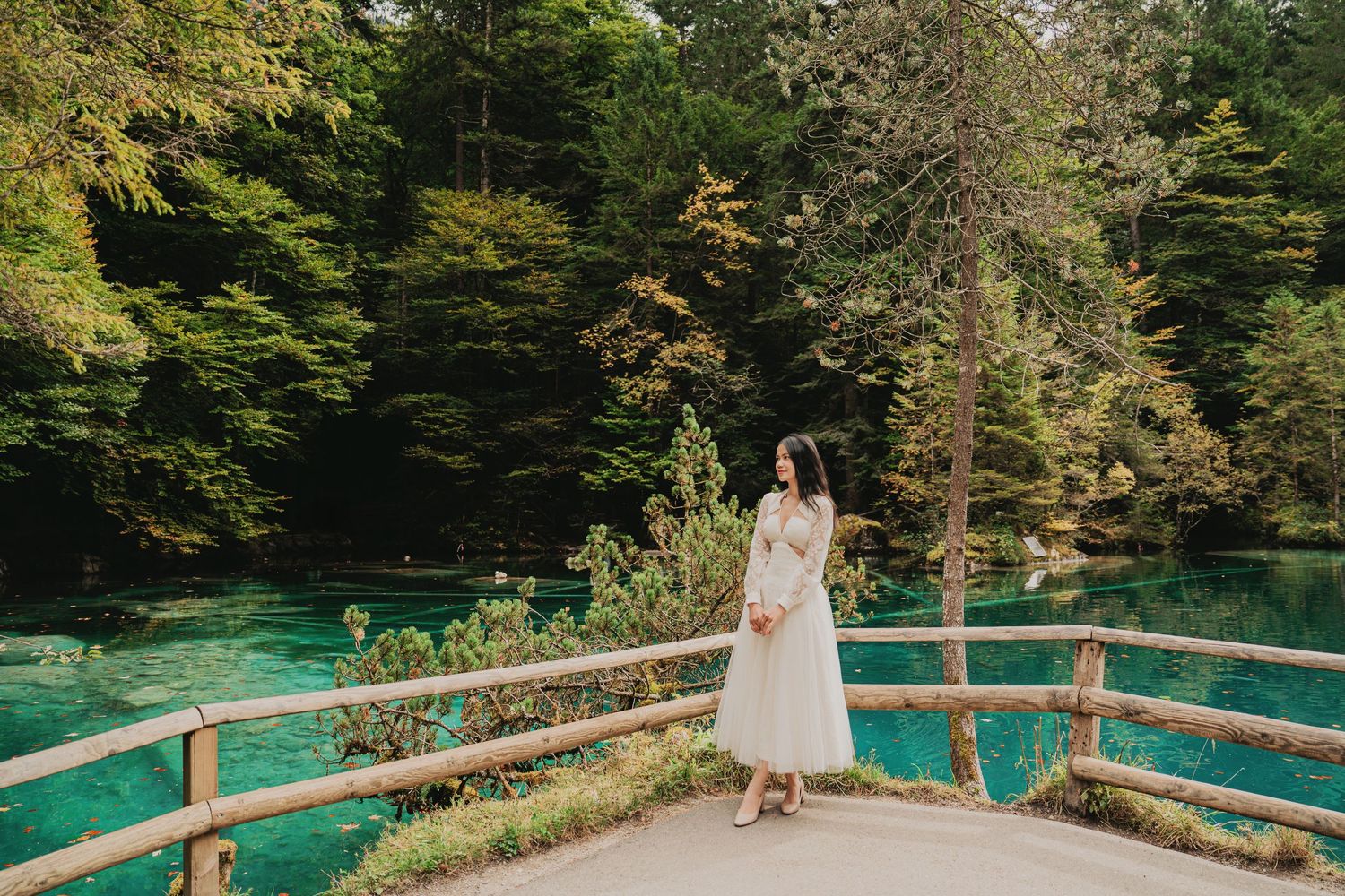 Woman in white midi dress stands on wooden bridge overlooking vivid turquoise alpine lake.