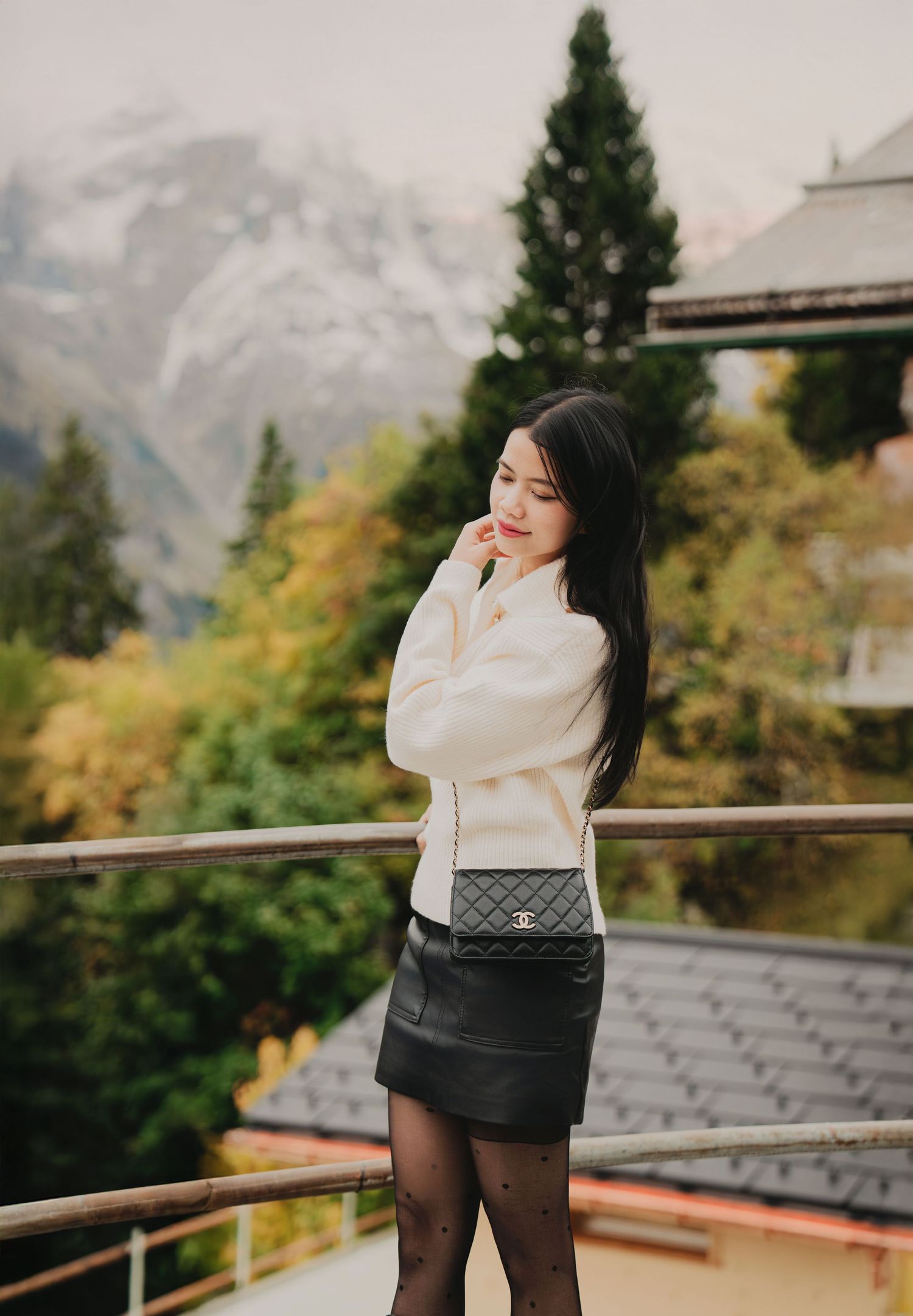 Young woman in cream sweater and black mini skirt poses with Chanel bag against Alpine mountain backdrop.