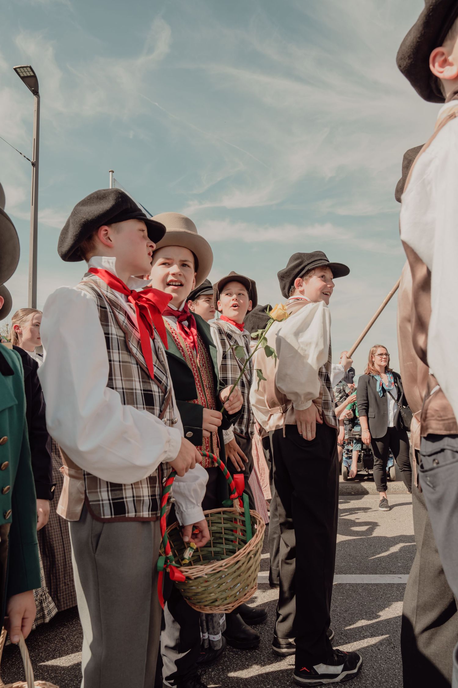 Boys in traditional folk costumes with hats and scarves carry flower basket at outdoor parade.