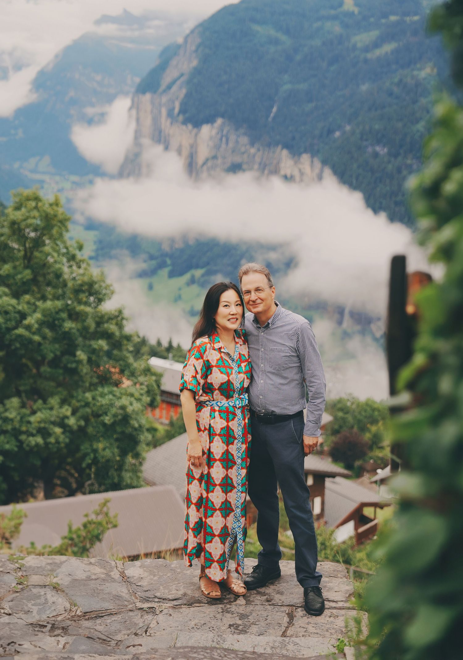 Smiling couple stands on stone terrace with misty Swiss Alpine valley and village rooftops behind them.