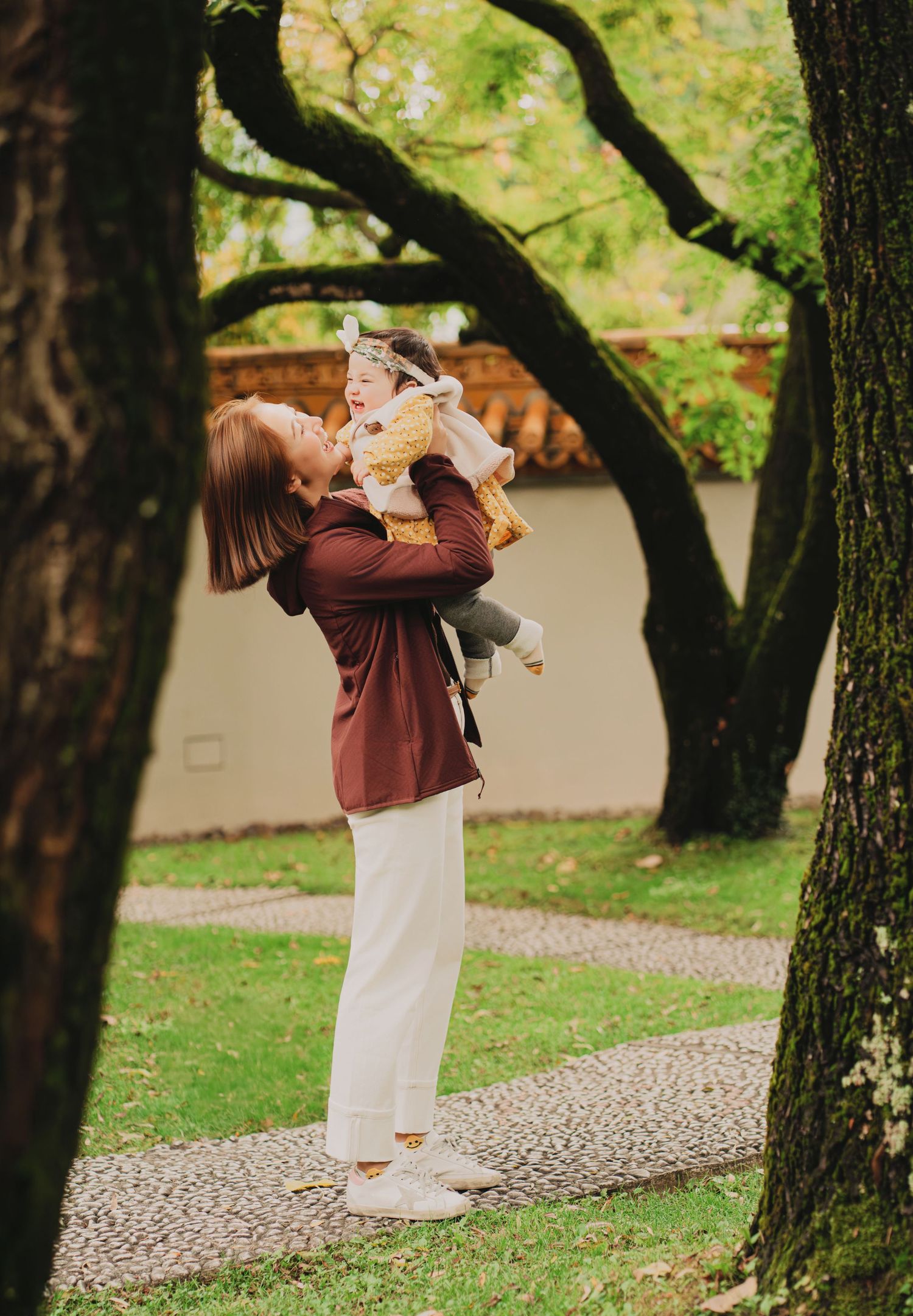 Mother lifts smiling baby overhead beneath large trees in a scenic garden pathway.