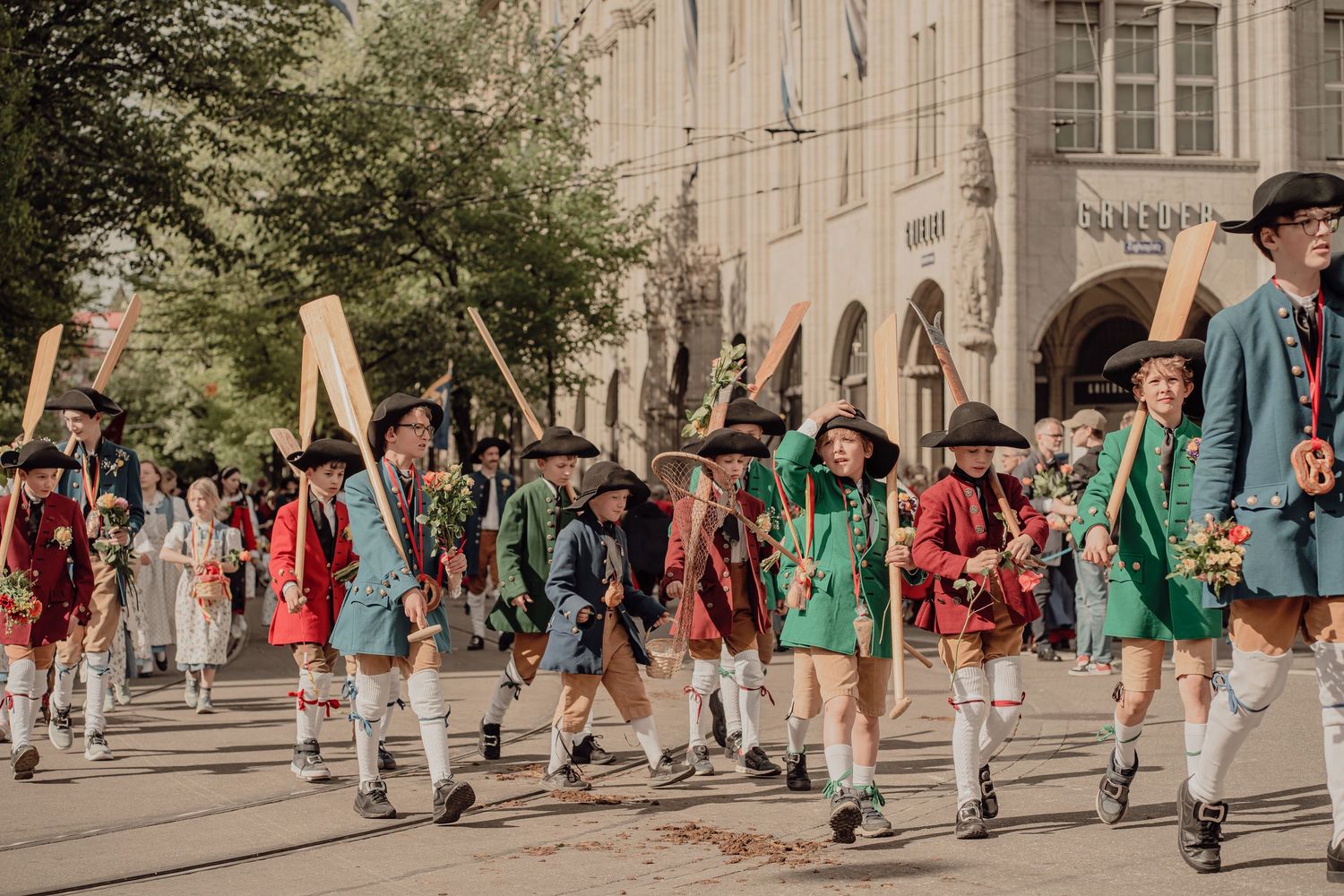 A large group in colorful 18th-century costumes marches with wooden poles in a sunny historical street parade.