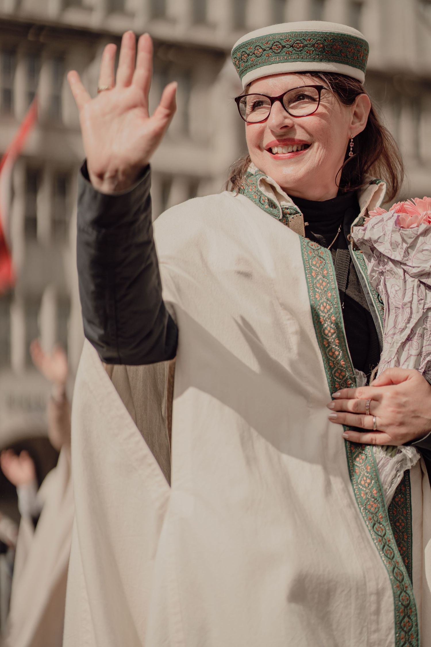 Smiling woman in white robe with green trim hat waves at traditional festival parade.