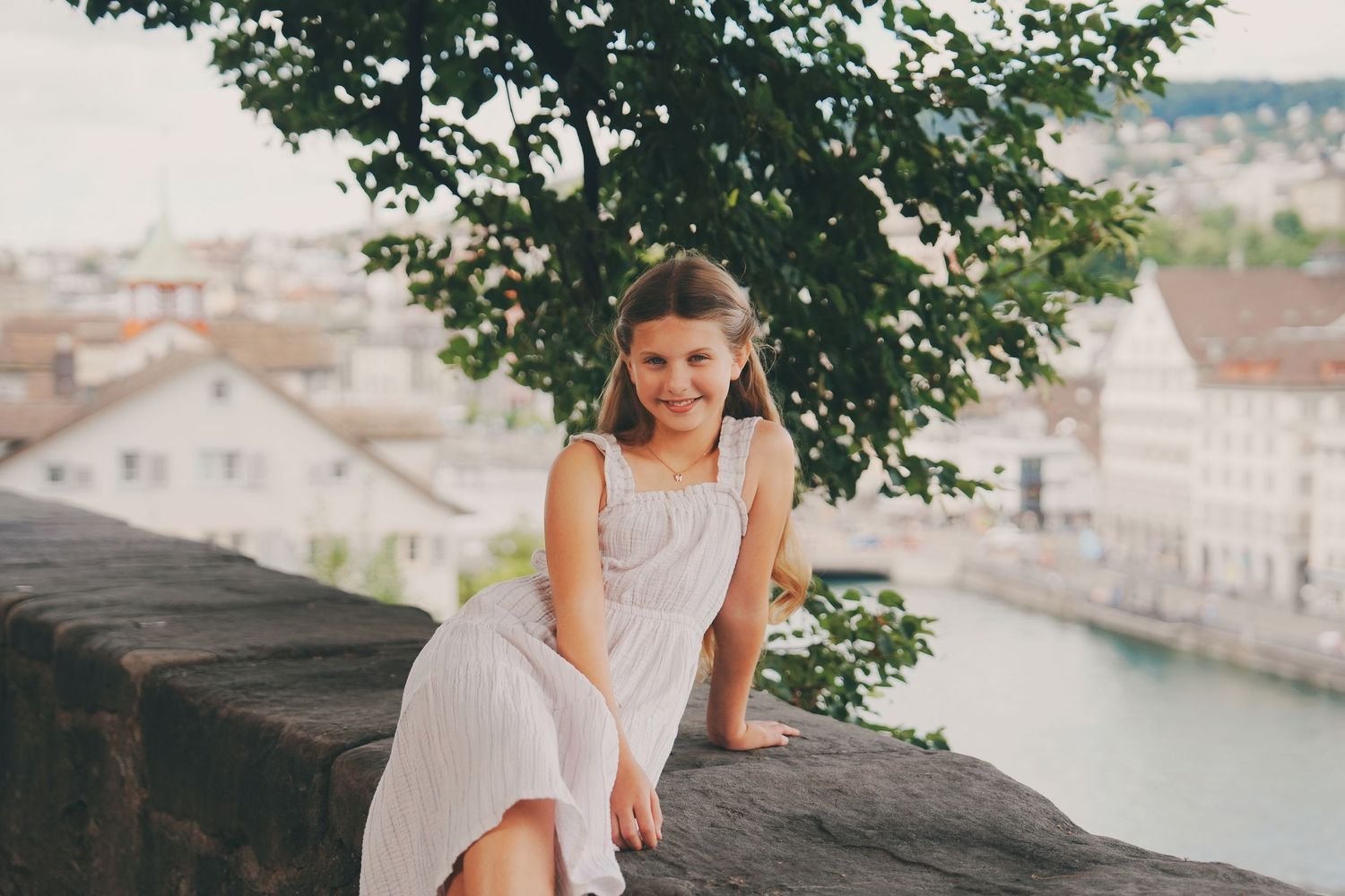 A smiling girl in a white dress sits on a stone wall with a European river city in the background.