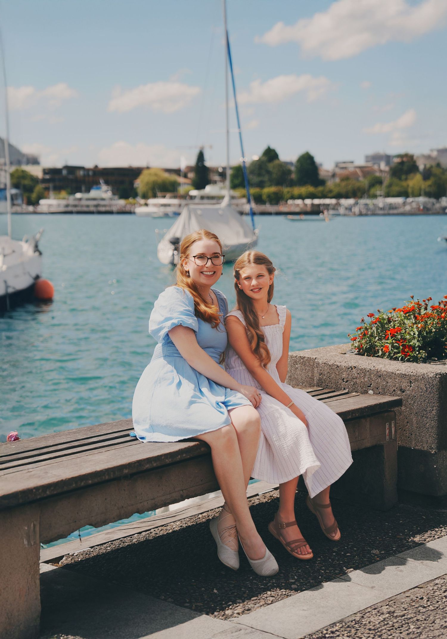 Woman and girl in summer dresses sitting on bench by turquoise lake with sailboats.