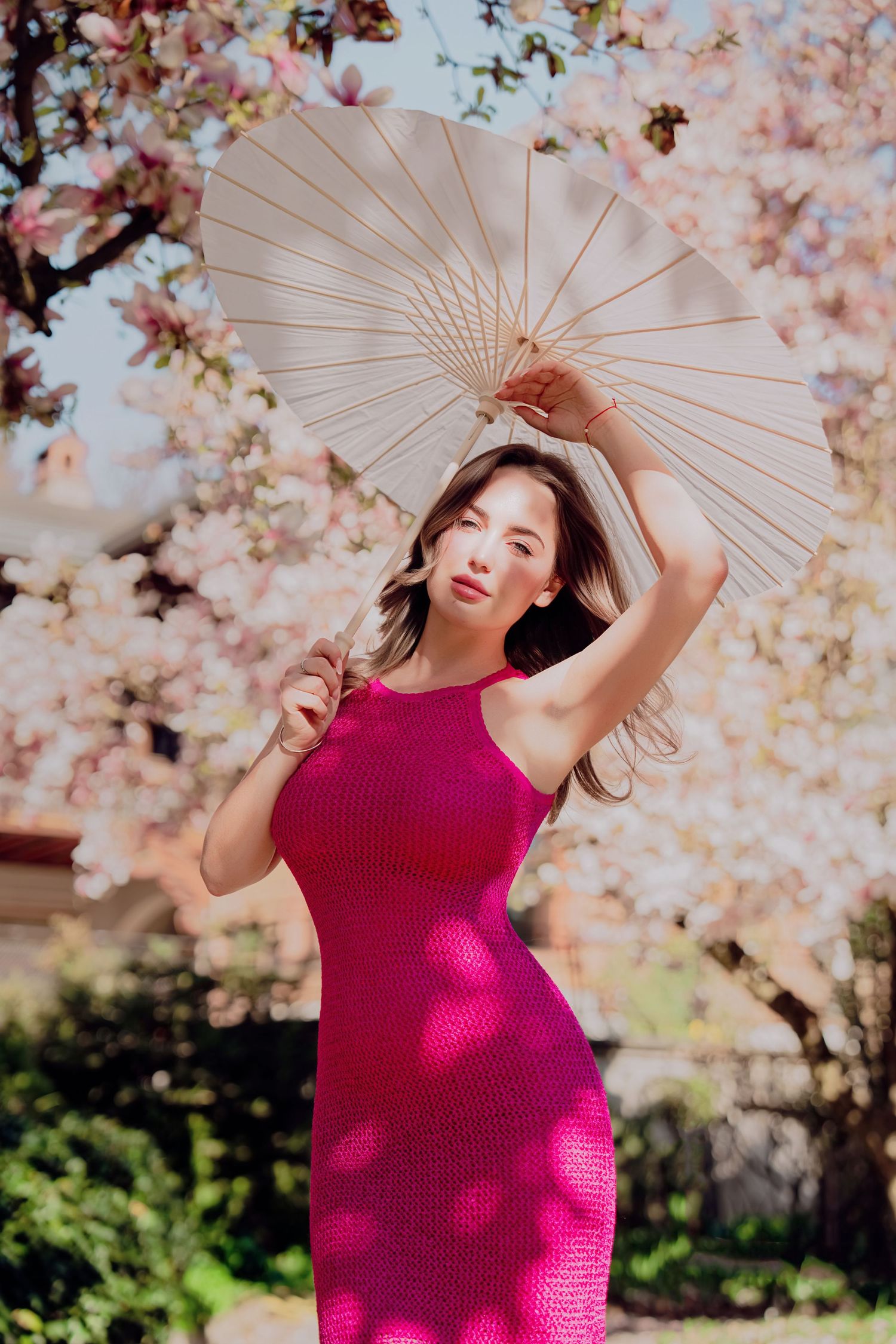 Woman in pink knit dress holding white parasol under blooming magnolia tree in sunlight.
