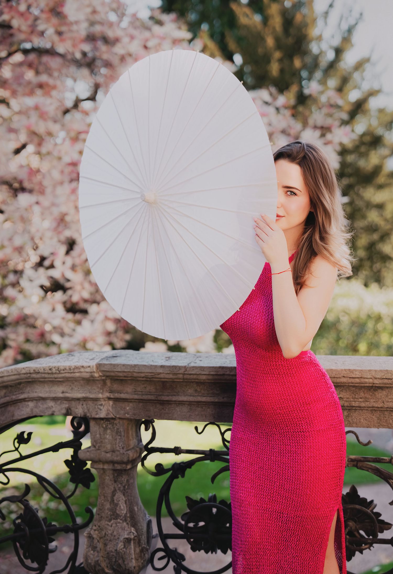 Woman in pink dress holding white parasol near stone balustrade with cherry blossoms.