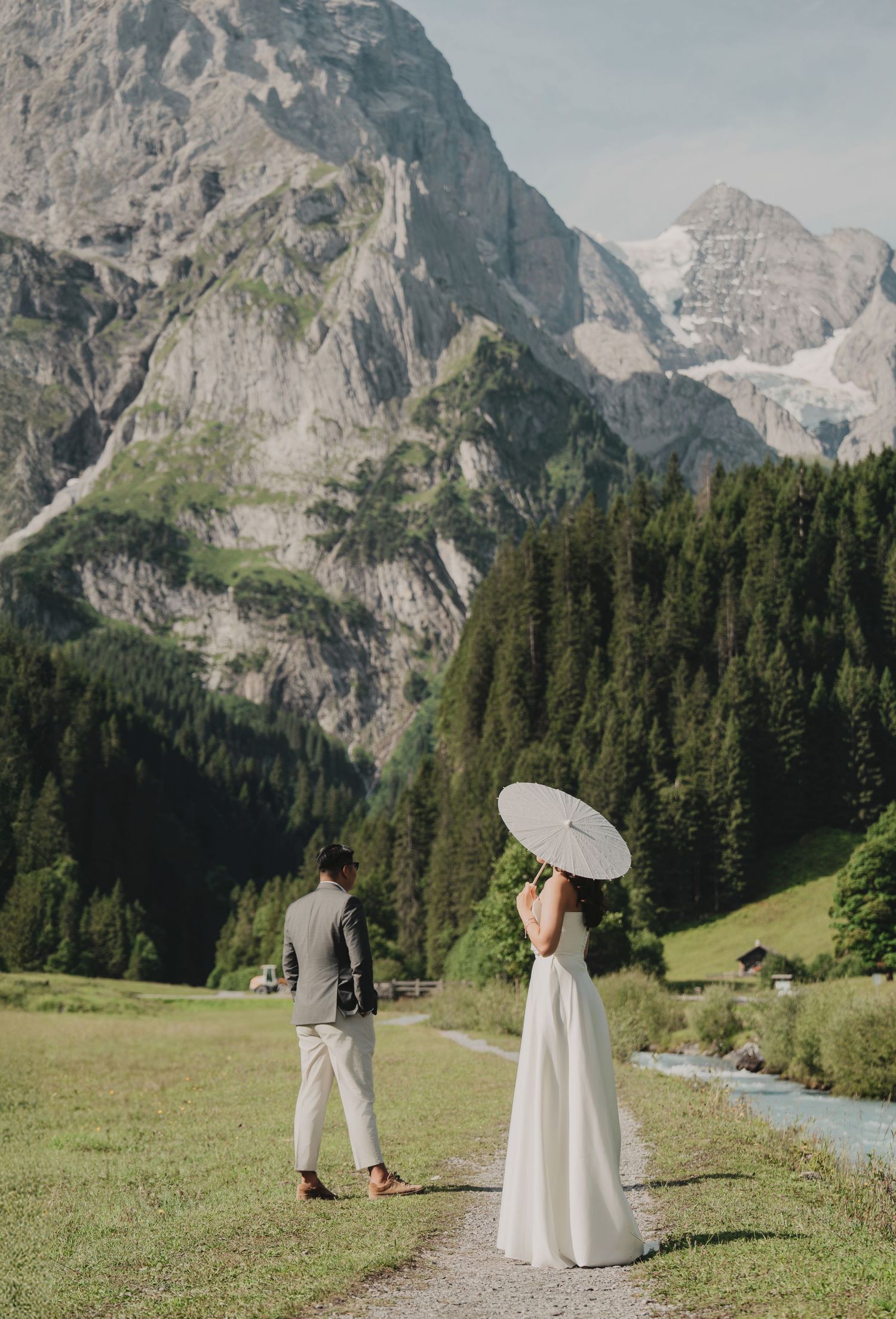 Bride holding white parasol facing groom on a path with towering rocky Alpine mountains behind.