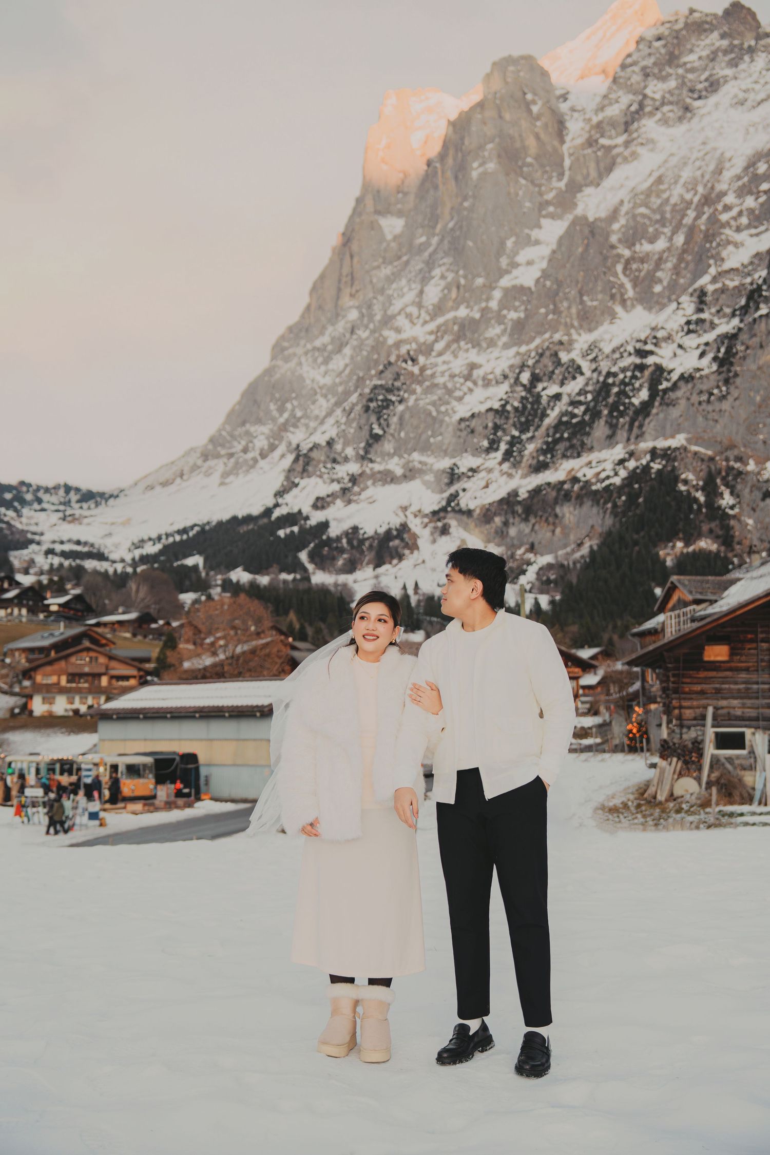 Couple in white winter outfits walking together in snowy alpine village with towering mountain at sunset.