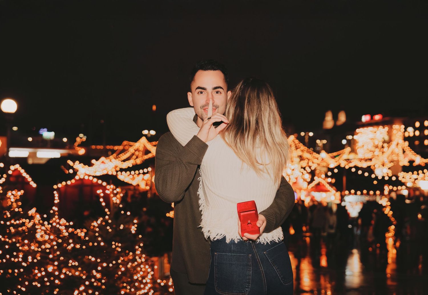 Happy couple embracing at a festive Christmas market surrounded by glowing golden holiday lights.