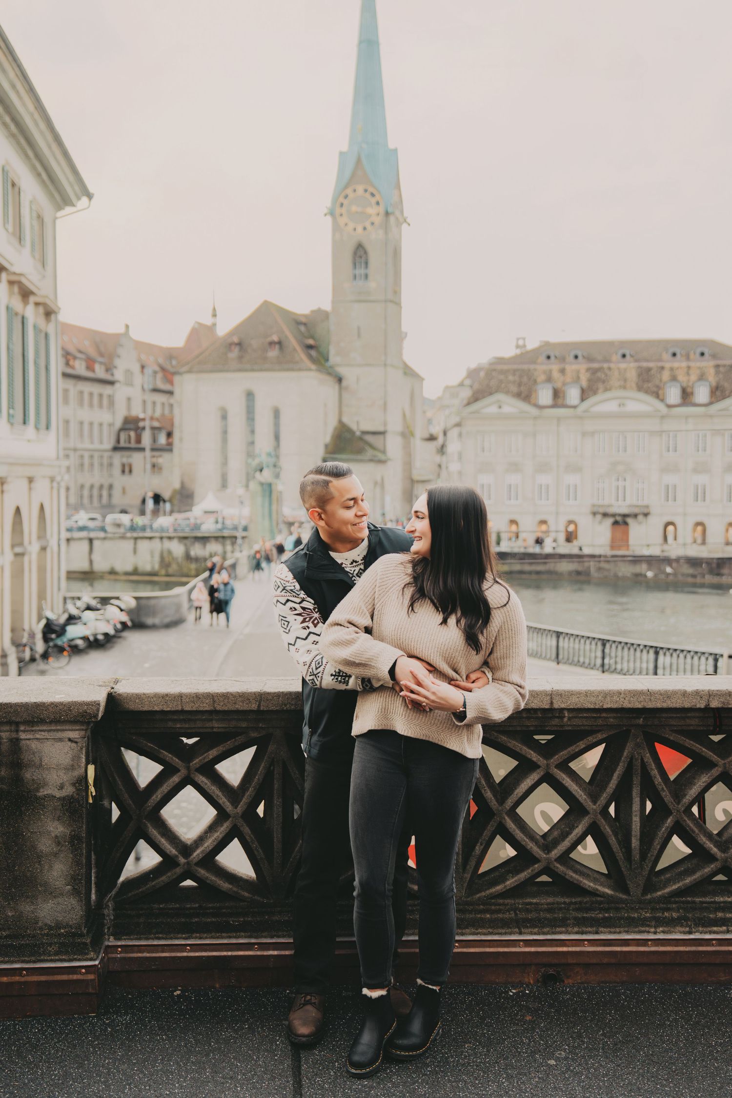 Couple standing close together on a bridge with Zurich old town and church tower behind them.