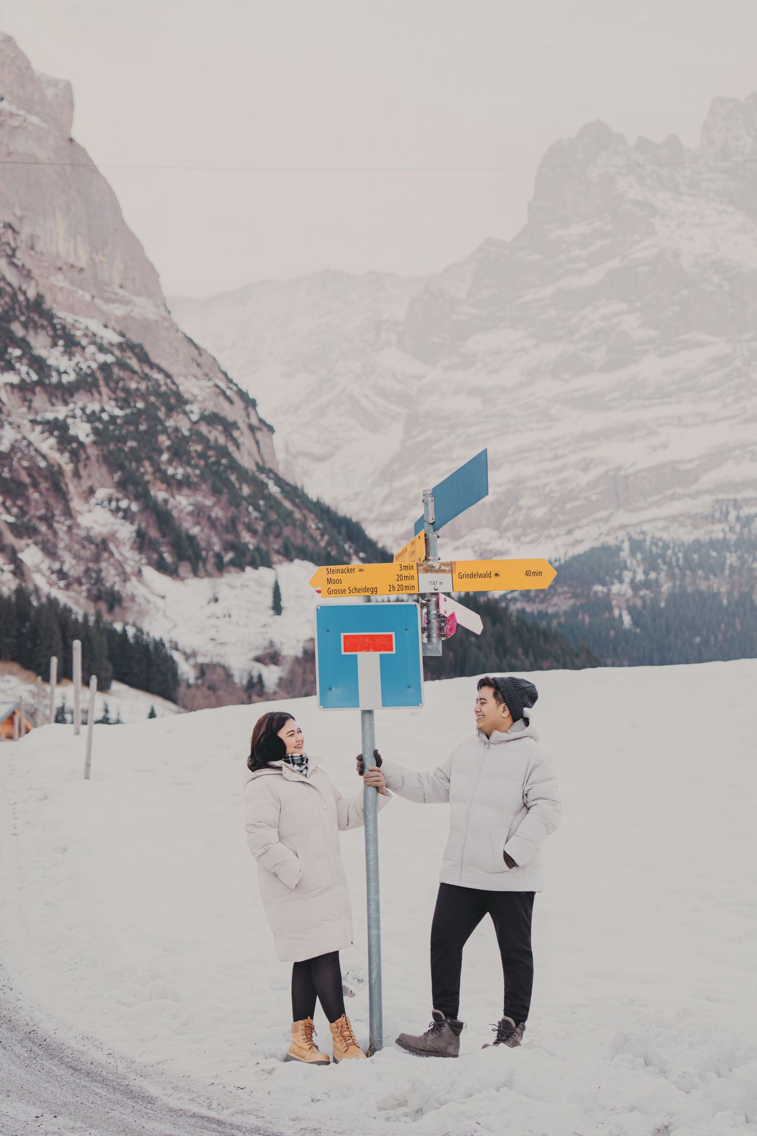 Couple in white coats at a colorful alpine signpost surrounded by snow-covered mountain landscape.