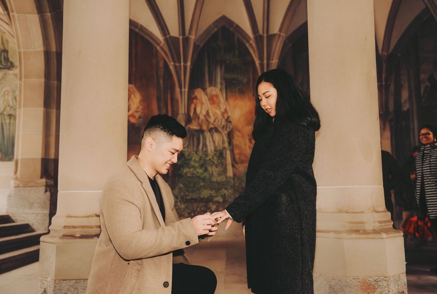 Man kneeling proposing with ring to woman inside gothic arched hall with painted murals.