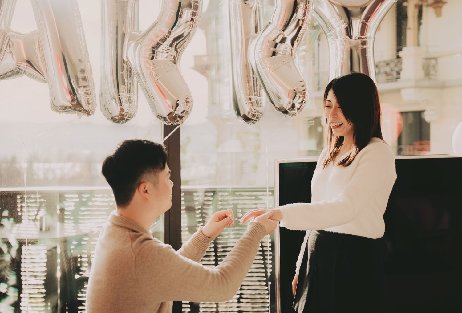 Man proposing to woman with ring under silver letter balloons indoors.