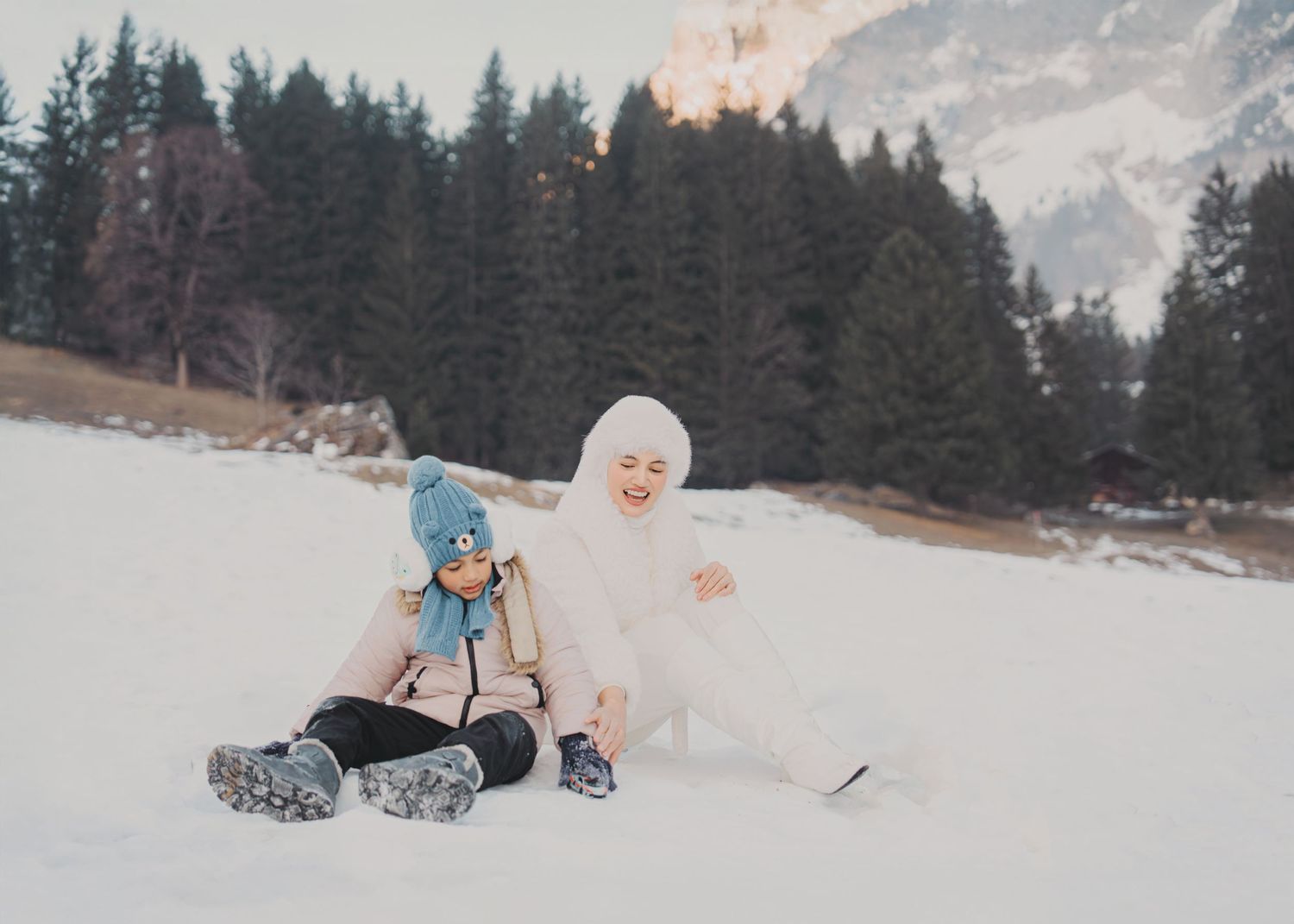 Mother and young daughter sitting and laughing together in snow with pine forest and mountains behind.