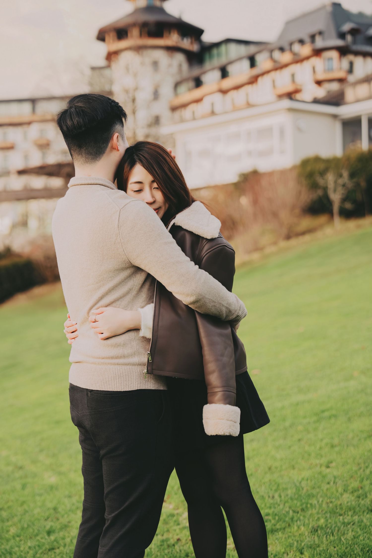 Asian couple sharing a warm embrace on green lawn outside luxury hotel building.