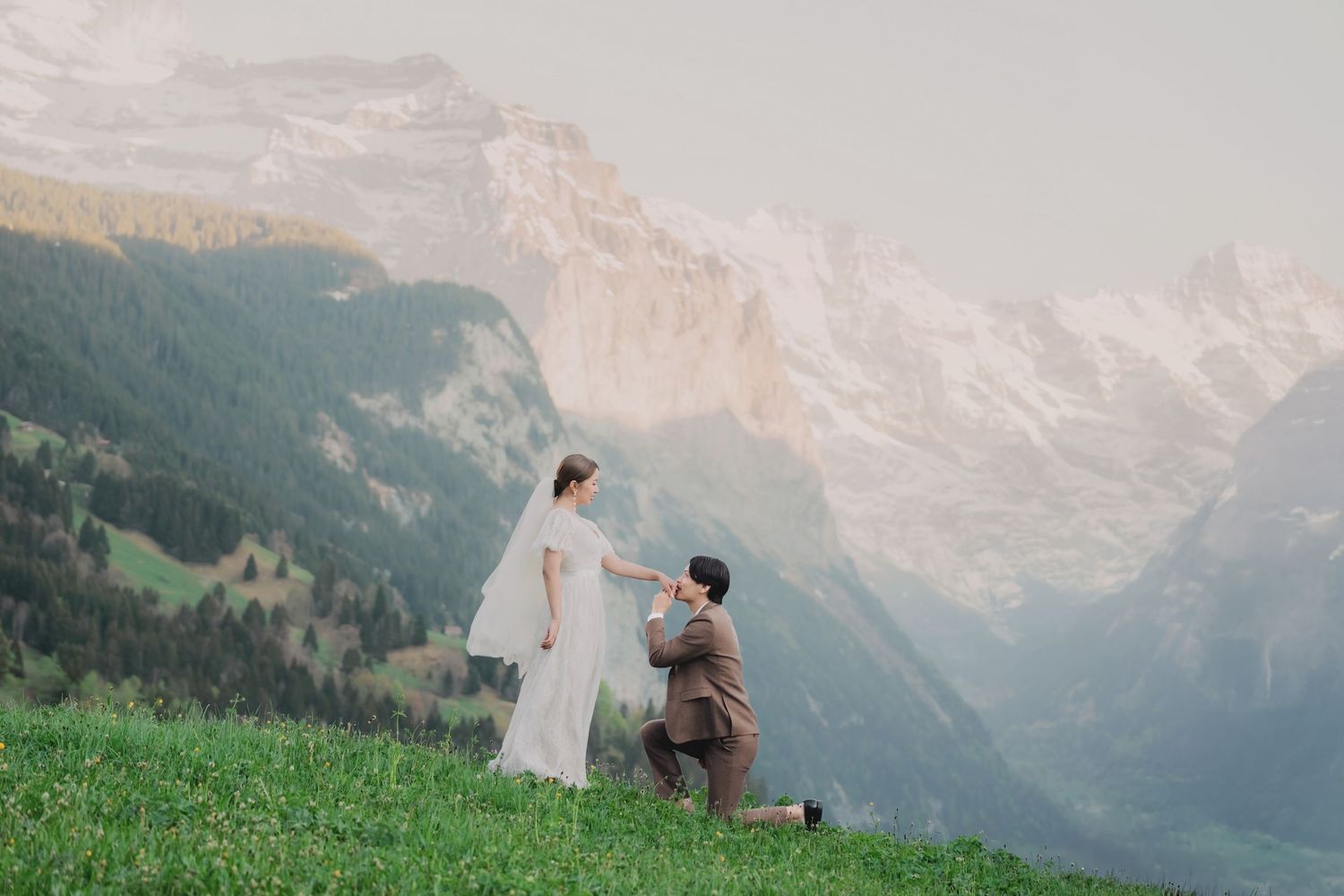 Groom kneeling before bride on lush alpine meadow with stunning Swiss mountain range and valley in background.